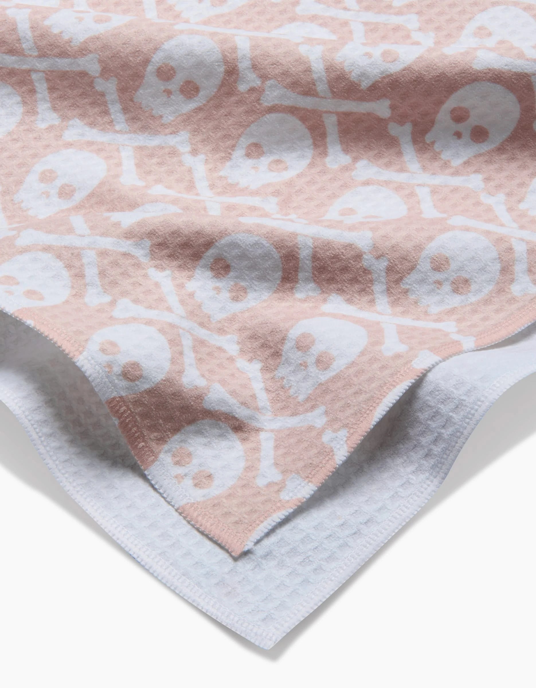 The Hufton Studio Skull Bones Natural Luxe Hand Towel, featuring a white skull and crossbones on soft pink, is layered over a white textured towel. Both ultra-absorbent towels add style and function to your bathroom decor.
