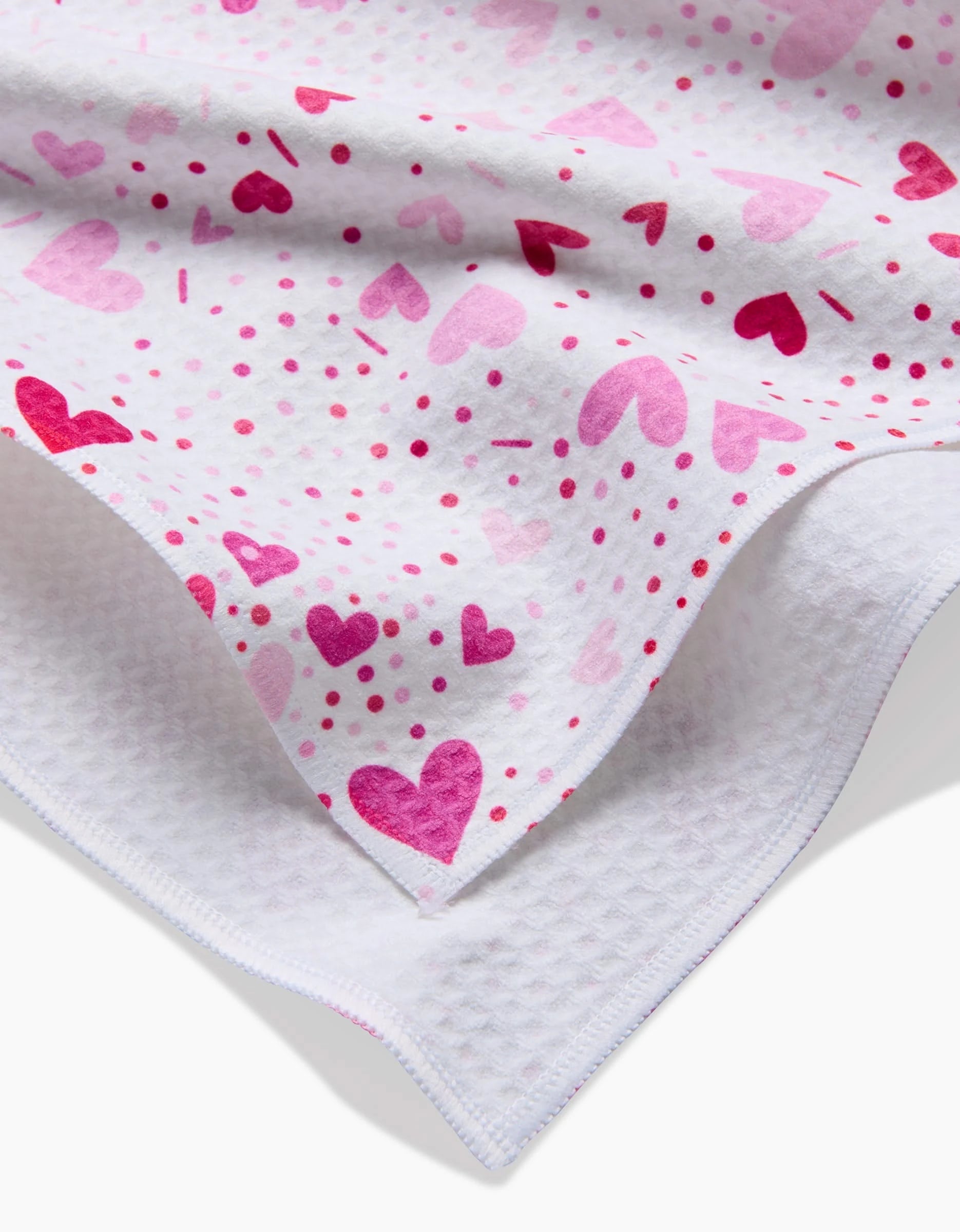 A close-up of the Sprinkled With Love Luxe Hand Towel by Rebecca Bobko, featuring pink and magenta heart and dot patterns on soft white fabric, ultra-absorbent and neatly folded over a textured white surface.