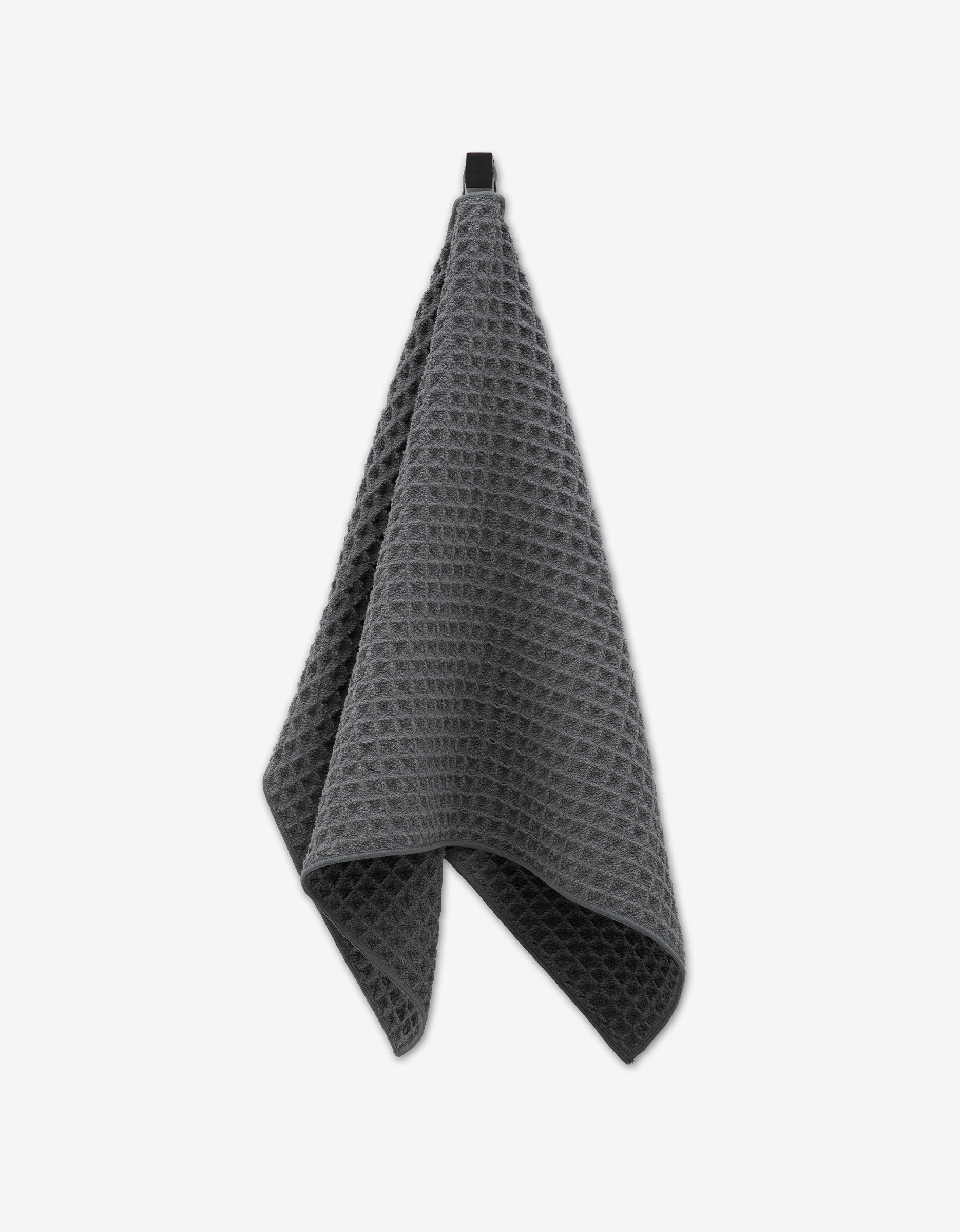 The GEOMETRY Charcoal Waffle Hand Towel, featuring a subtle texture, hangs from a black loop against a white background—offering eco-friendly luxury and modern style.