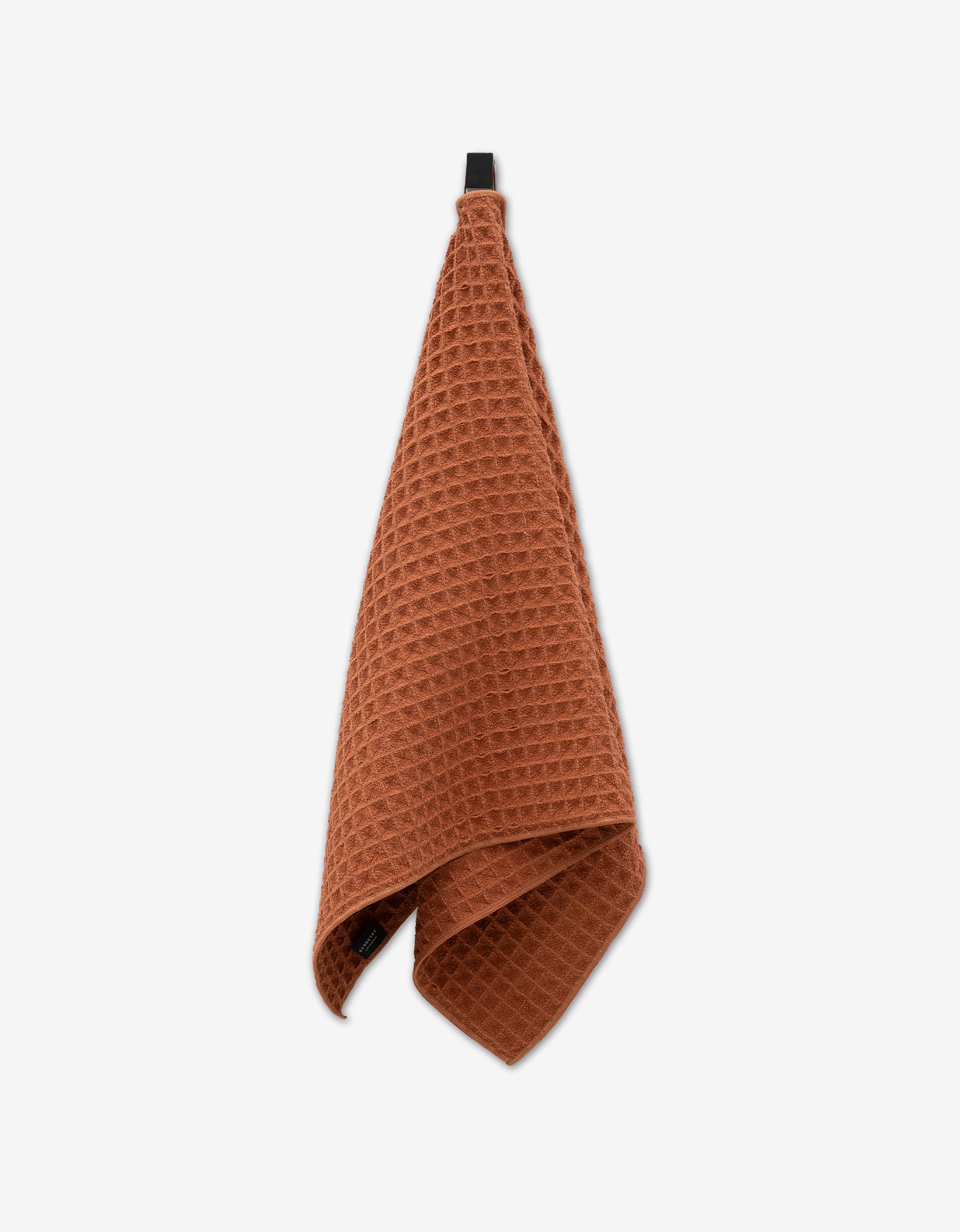 The GEOMETRY Clay waffle hand towel in a rust hue hangs from a black loop against a white backdrop, adding a touch of luxury to your space.