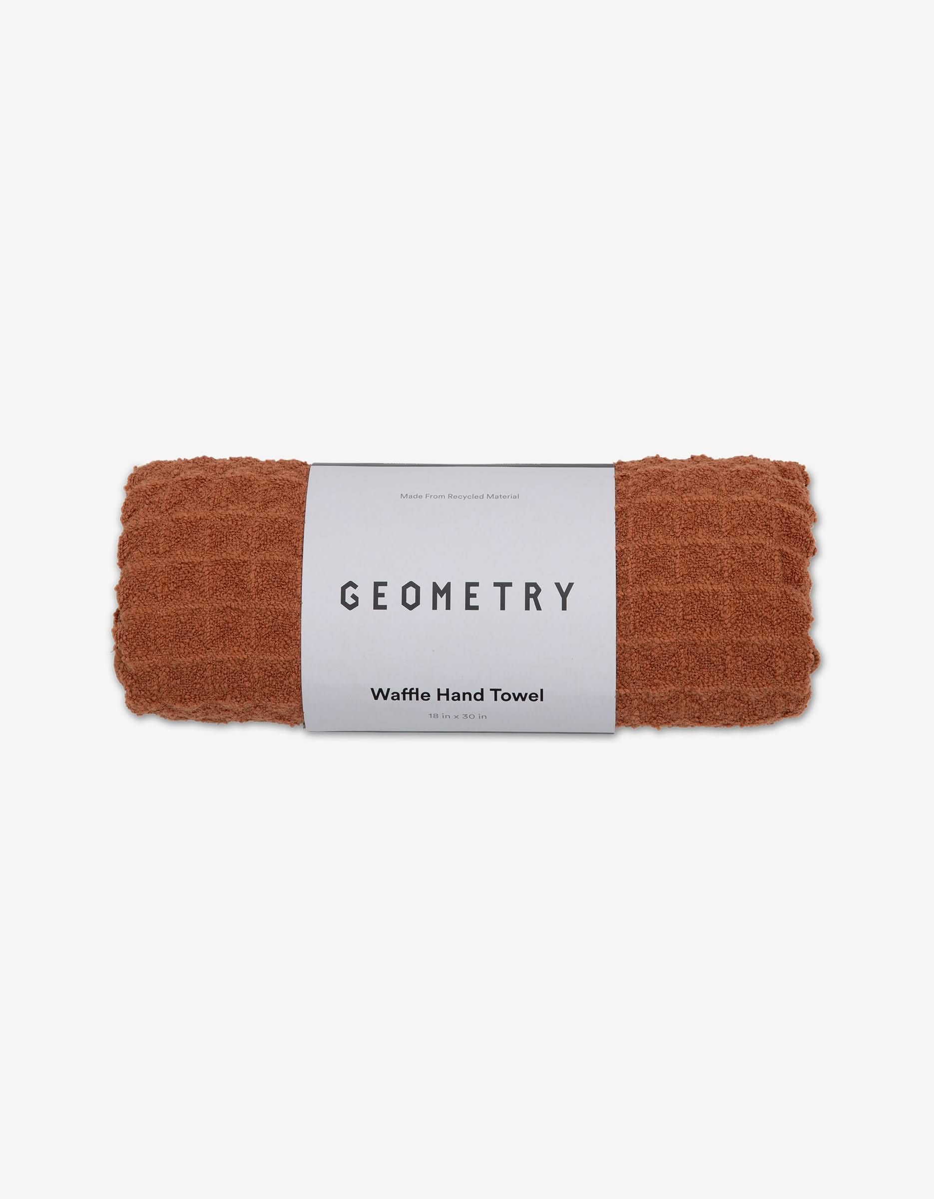 A rolled, rust-colored GEOMETRY Waffle Hand Towel in the shade Clay with a white label sits on a plain white background—an eco-friendly towel that adds luxury to your space.