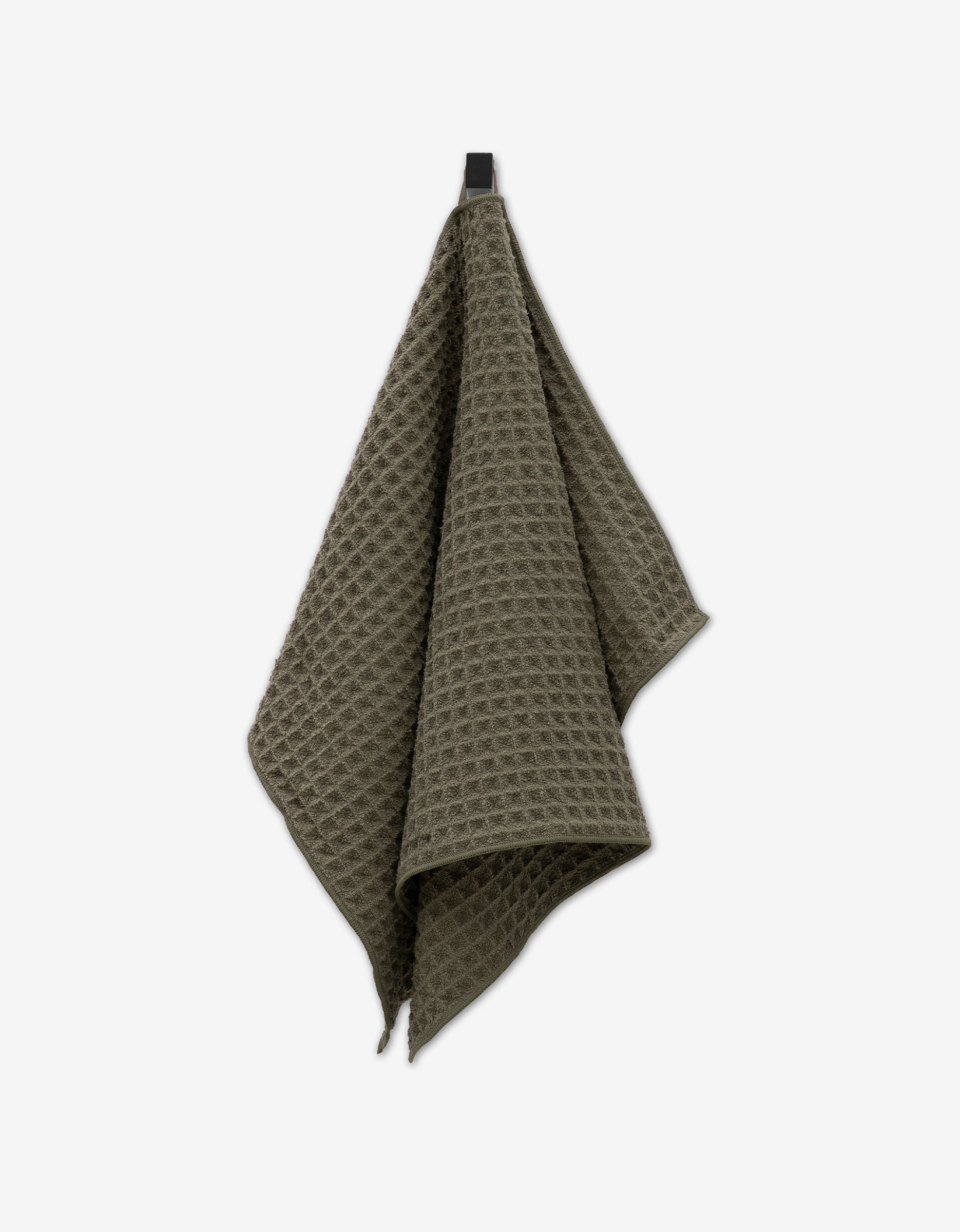 A GEOMETRY Forest Waffle Hand Towel in olive green hangs from a black hook against a plain white background, showing off its textured waffle pattern.
