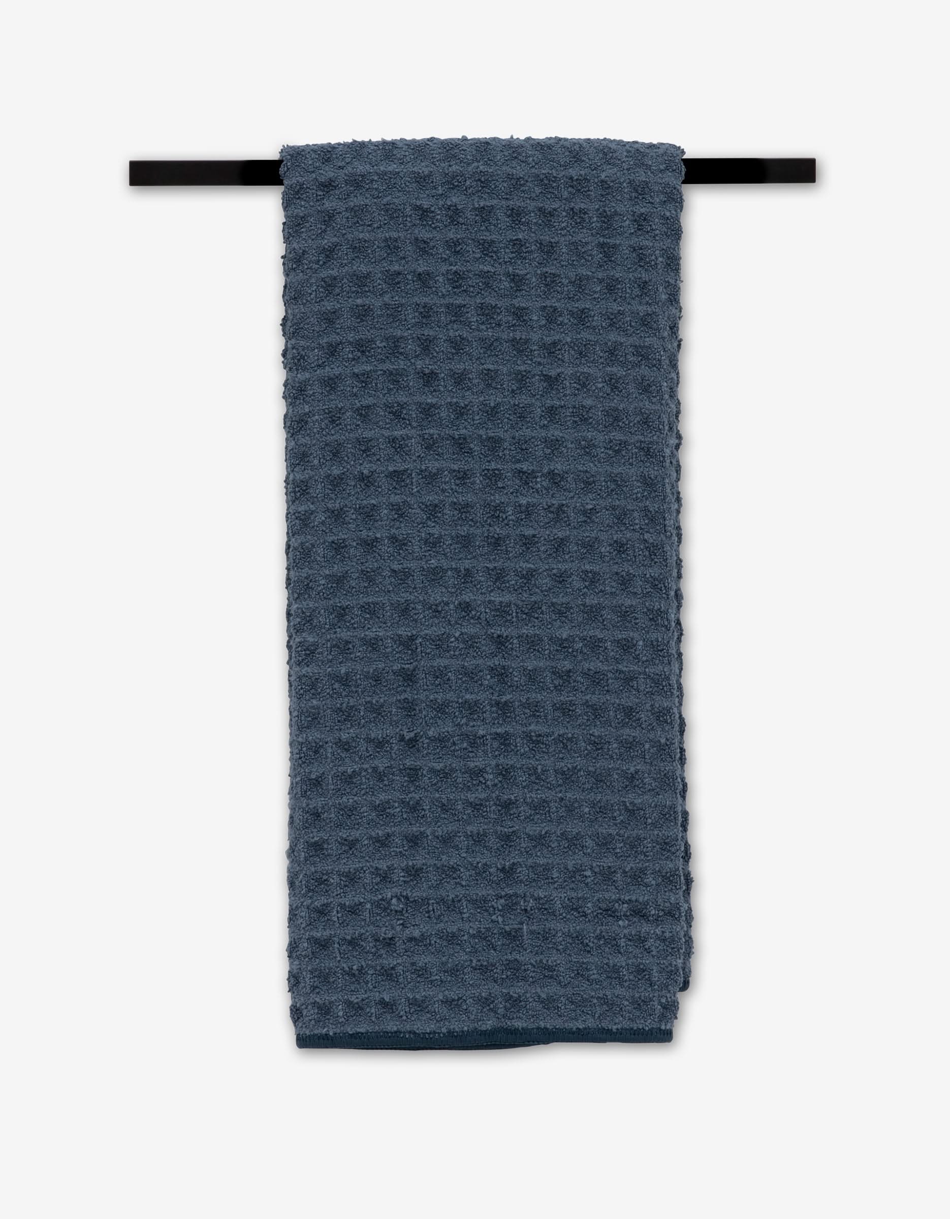 A Midnight Blue Waffle Hand Towel by GEOMETRY, made from recycled materials, is neatly folded over a black towel bar against a white background.