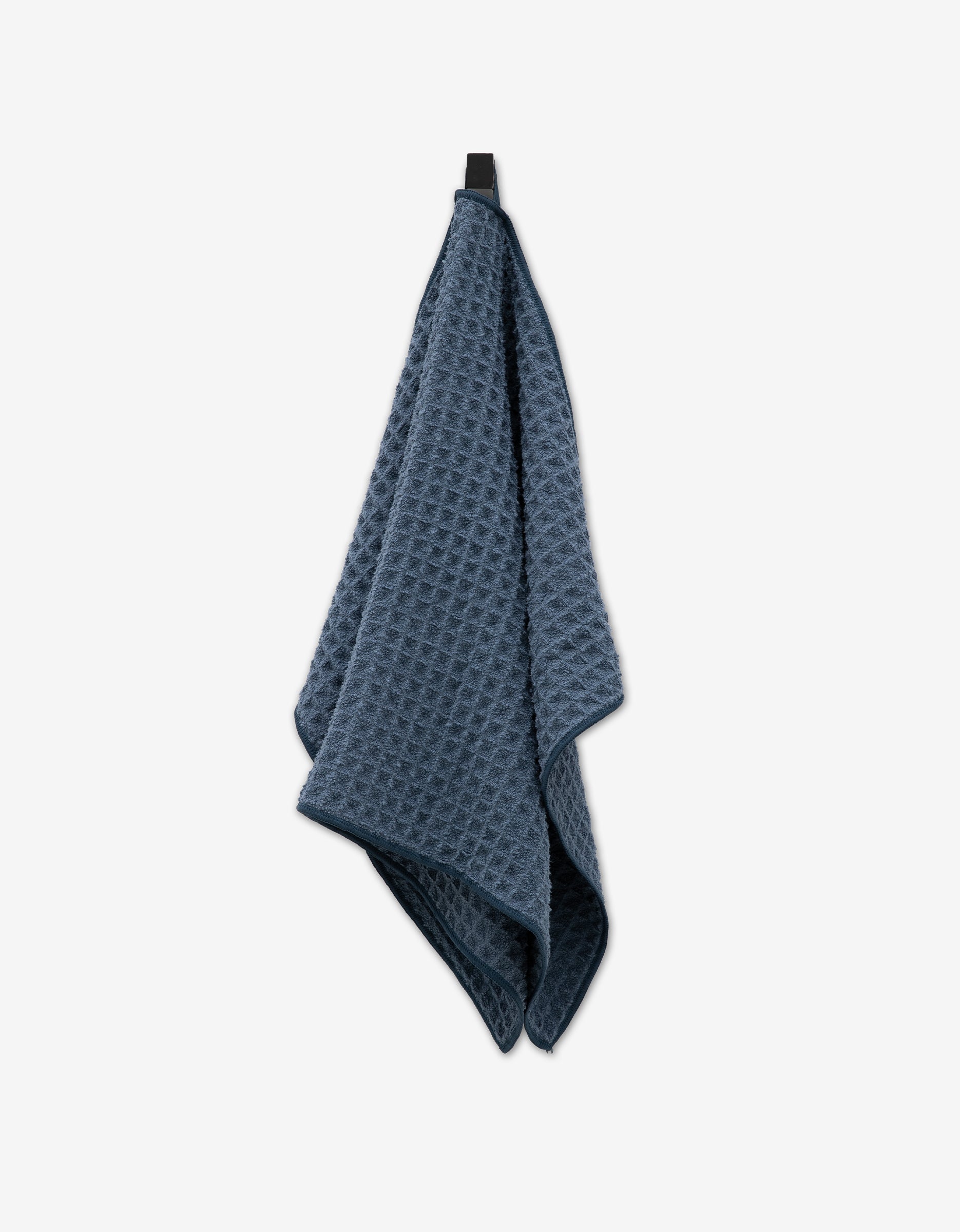 The GEOMETRY Midnight Blue Waffle Hand Towel, with its rich texture, hangs gracefully from a black hook on a plain white background, exemplifying understated everyday elegance.