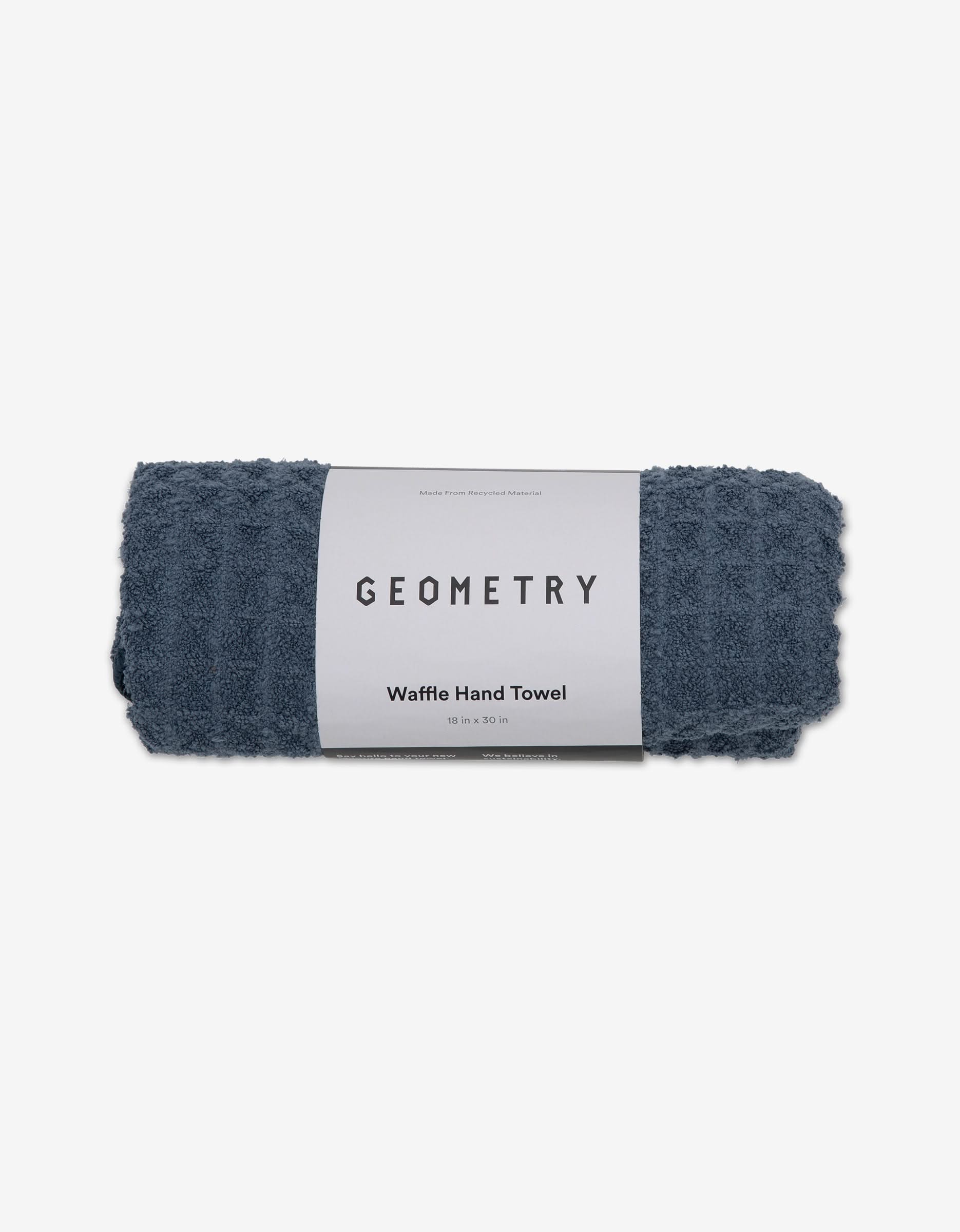 A rolled-up Midnight Blue Waffle Hand Towel by GEOMETRY, made from recycled materials and wrapped with a branded label, is displayed on a plain white background.
