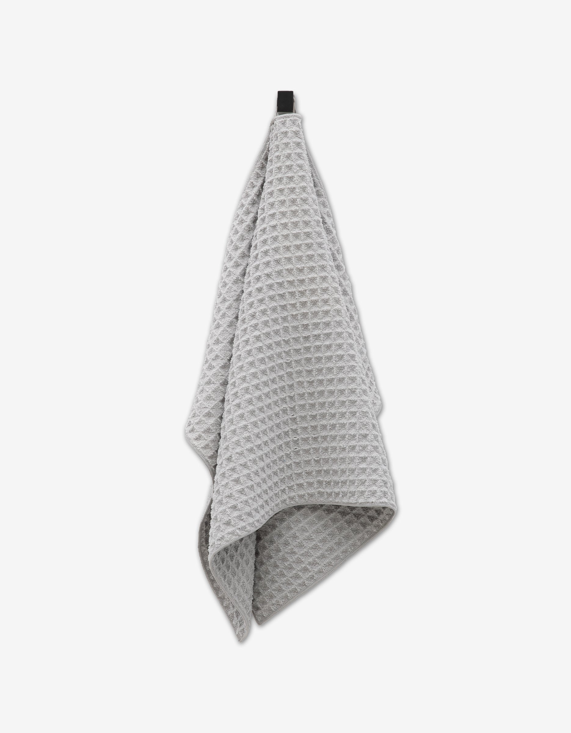 The GEOMETRY Stone towel, featuring a light gray waffle pattern, hangs from a black hook against a white background, highlighting its ultra-absorbent design.