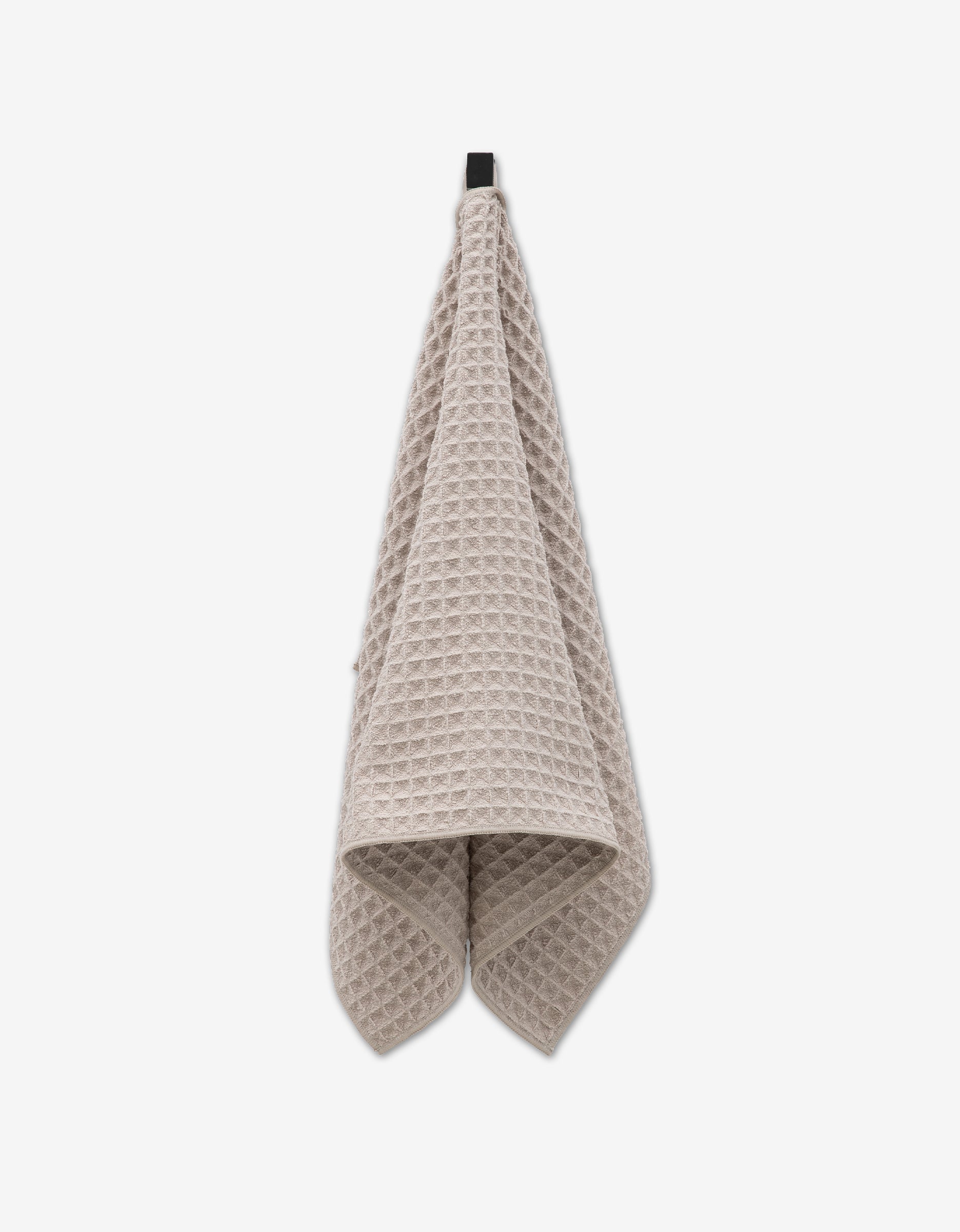 A taupe Waffle Hand Towel from GEOMETRY, made from recycled materials, hangs from a black hook against a plain white background.