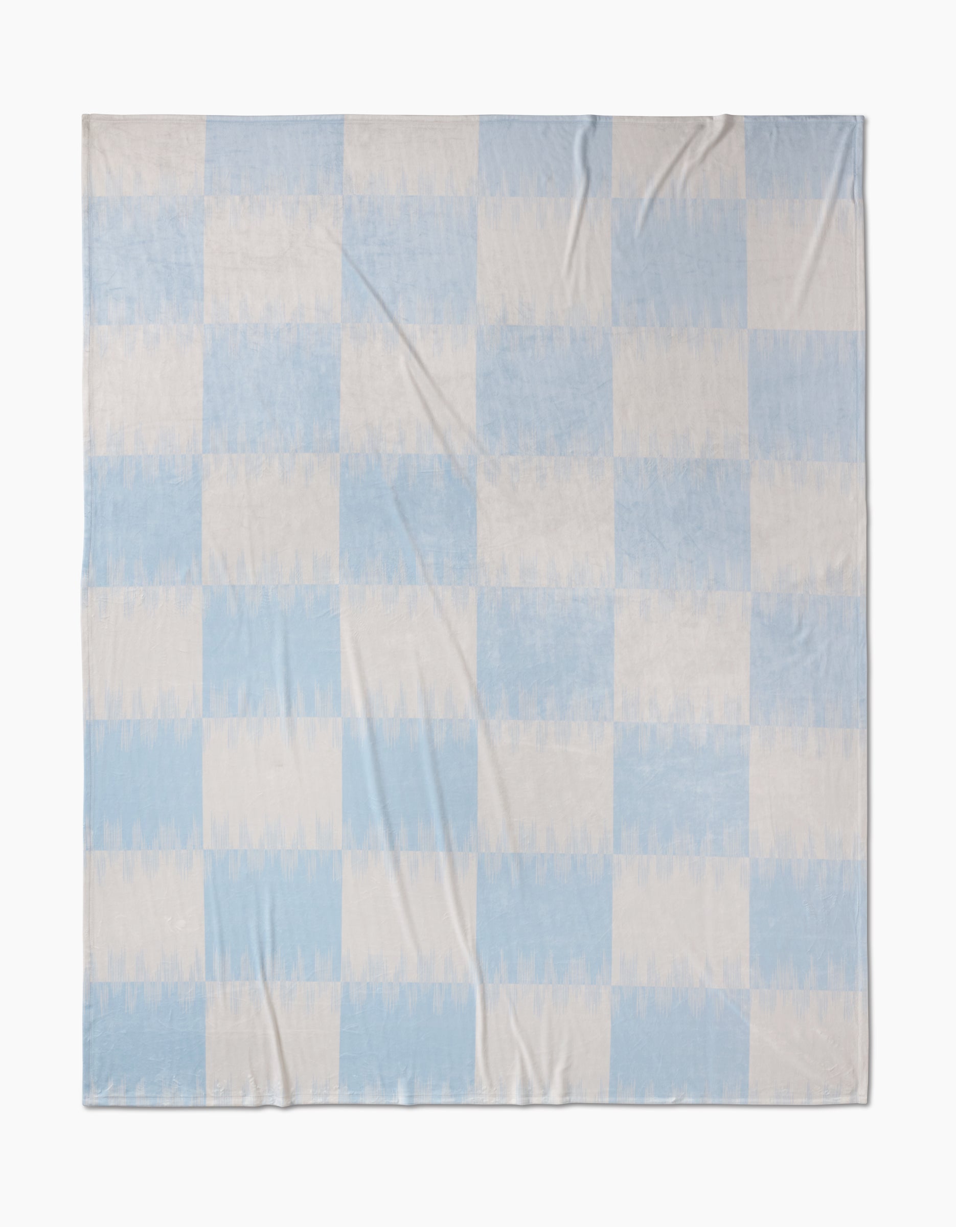 The Geometry Sweet Checks Butter Blanket showcases a simple checkered pattern with light blue and off-white squares, crafted from recycled materials. It’s laid flat, highlighting its soft fabric folds.