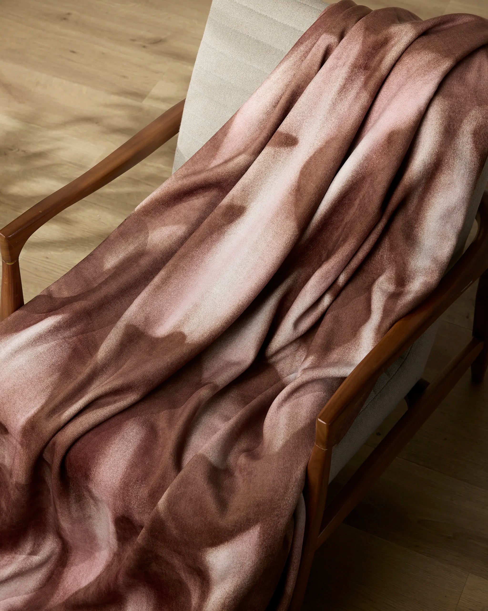 The Geometry Annie throw blanket, featuring brown and pink marbled patterns, is draped over a light beige cushioned wooden chair on a light wood floor.