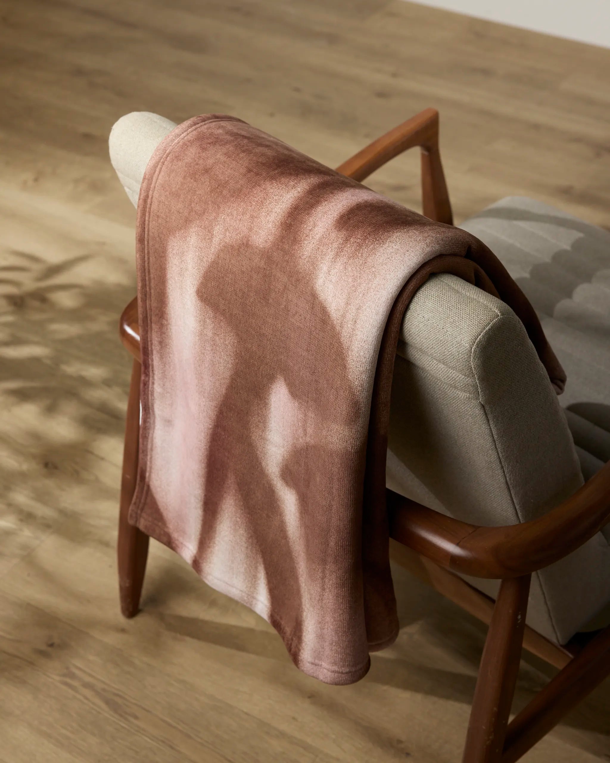 The Annie chair by Geometry, featuring beige upholstery and wooden construction, sits in a warmly lit room with a soft throw blanket draped over its backrest, casting a gentle shadow on the wooden floor.
