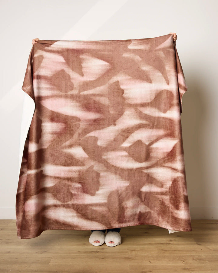 A person stands against a wall holding up the Annie blanket by Geometry, featuring a pink and brown pattern and made from recycled materials; only their feet in white socks peek out beneath the ultra-soft throw.