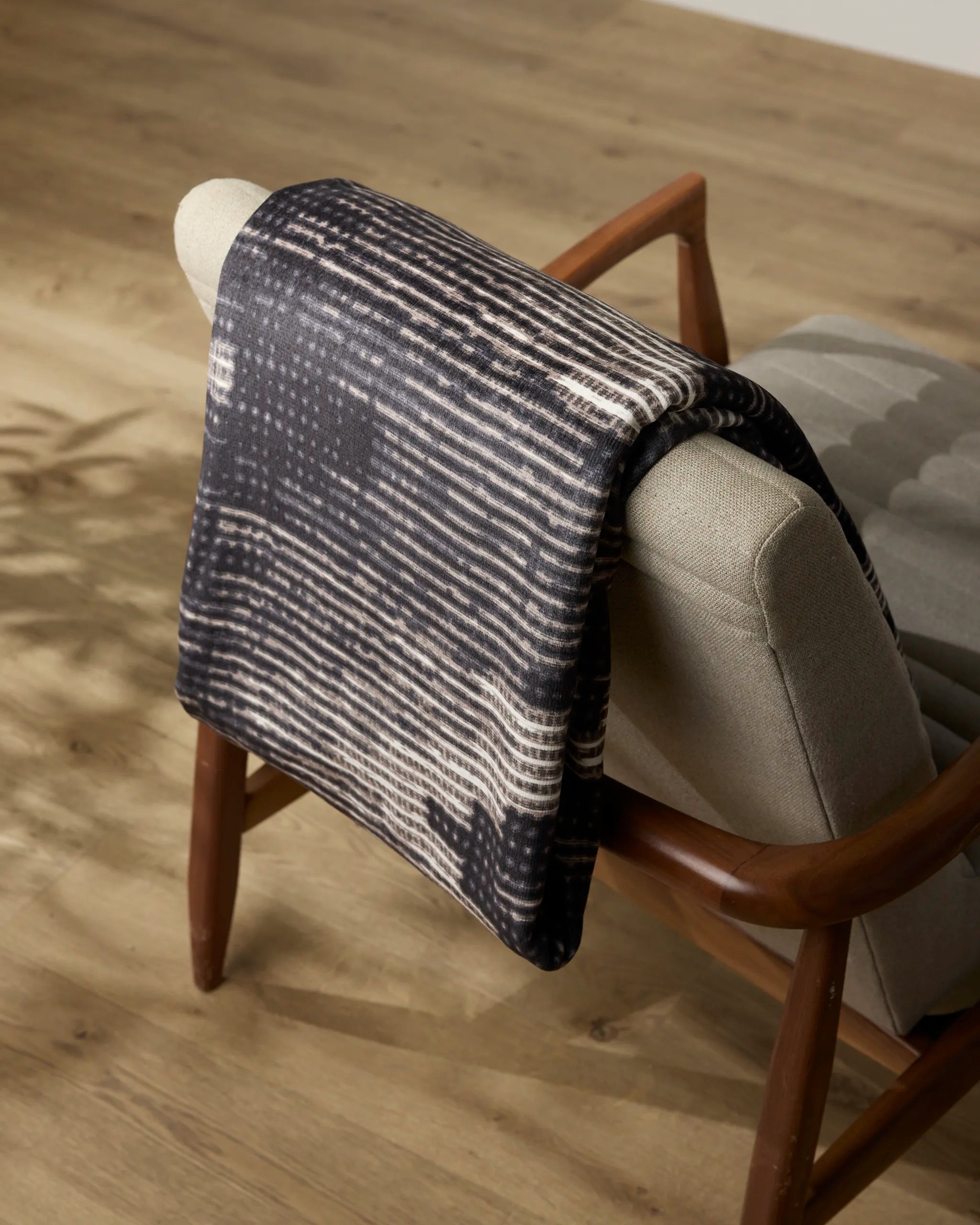 The Geometry Bowie throw blanket, featuring a black and white pattern, is draped over a mid-century modern armchair with wooden arms and light gray upholstery on a light wood floor.