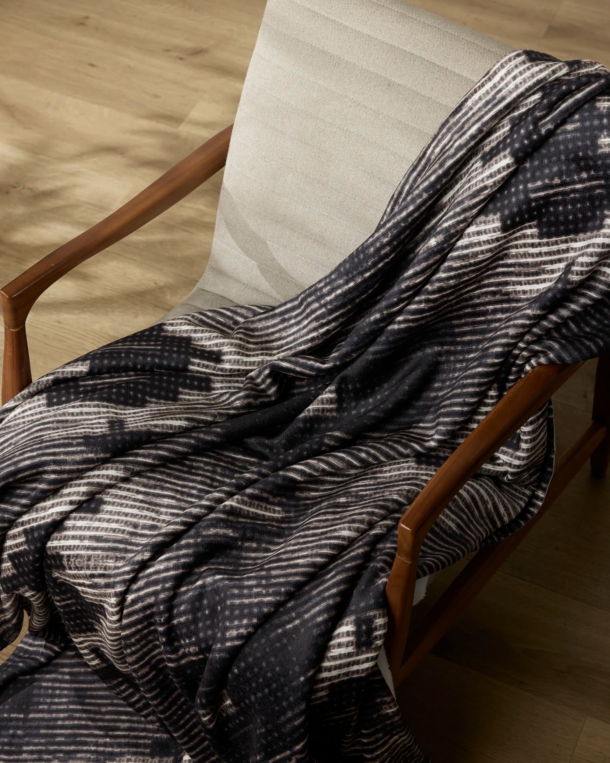 The Geometry Bowie wooden chair features a light fabric seat and backrest, draped with a soft throw blanket displaying white geometric patterns. Sunlight casts gentle shadows on the light wood floor beneath the chair.