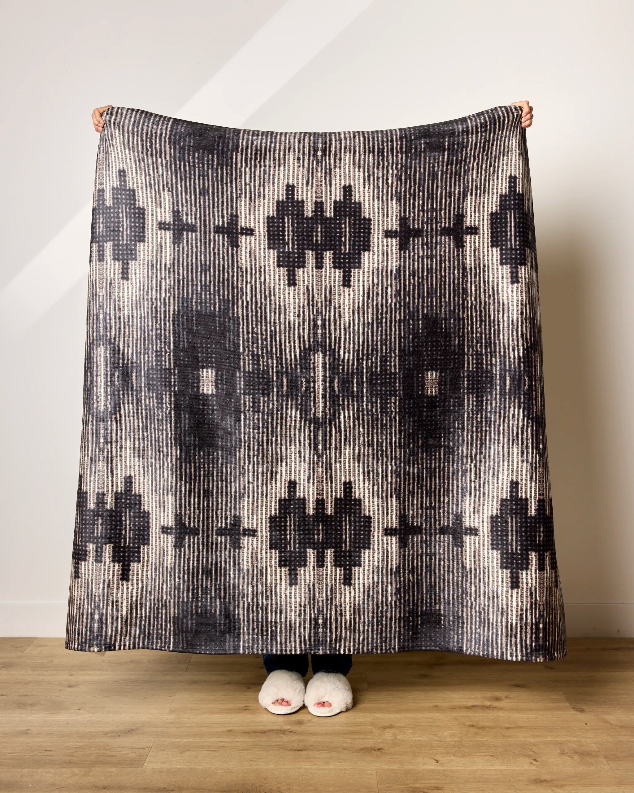 A person stands on a wooden floor holding up the ultra-soft Bowie blanket by Geometry in black and beige patterns, with only their hands and feet in white slippers visible behind it.