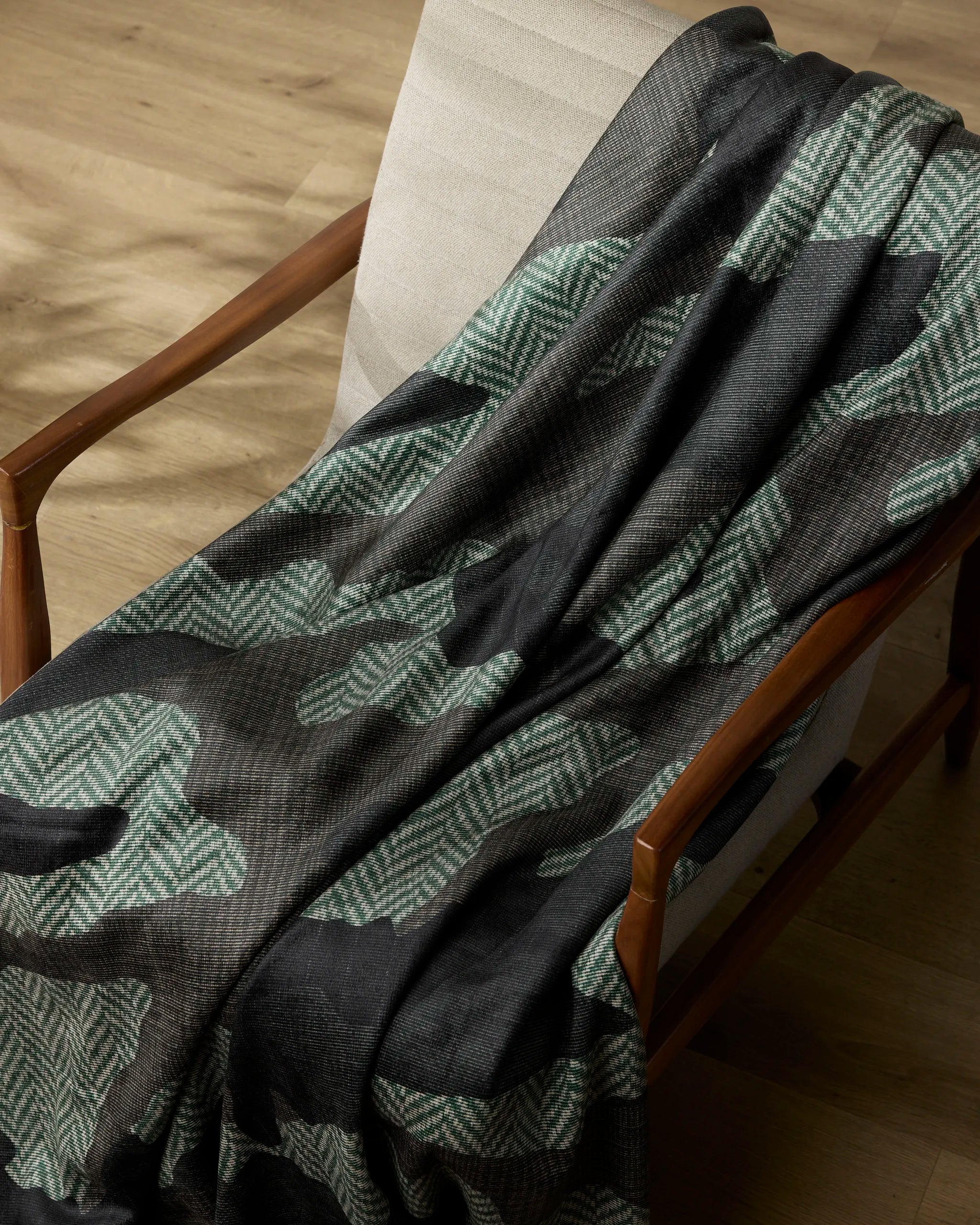 The Geometry Calloway blanket, featuring a black and green camouflage pattern, is draped over the armrest and seat of a light beige cushioned chair with wooden arms on a light wood floor.