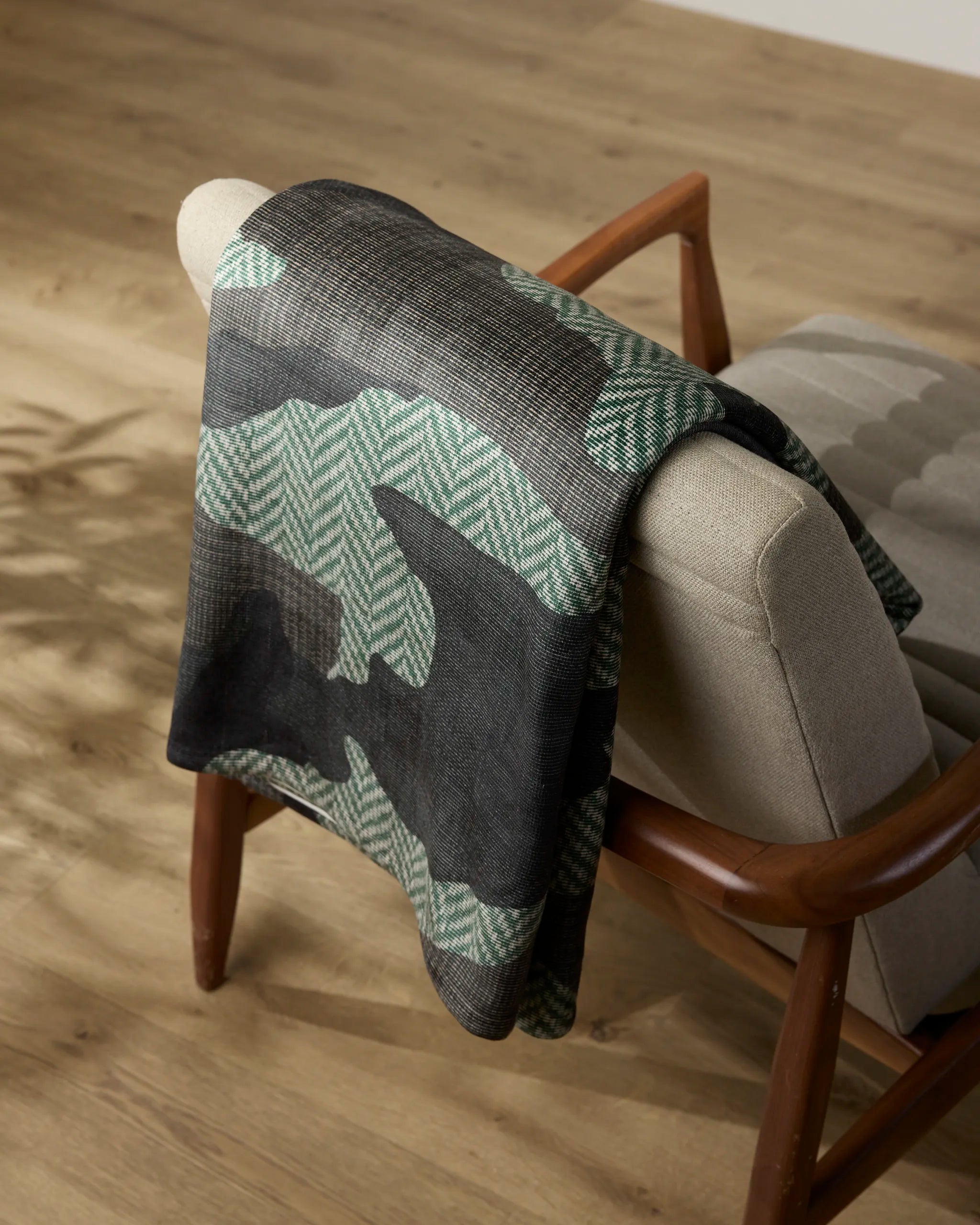 A soft, silky green and black Calloway blanket by Geometry is draped over a light beige upholstered chair with wooden arms on a light wood floor. Leaf shadows fall nearby.