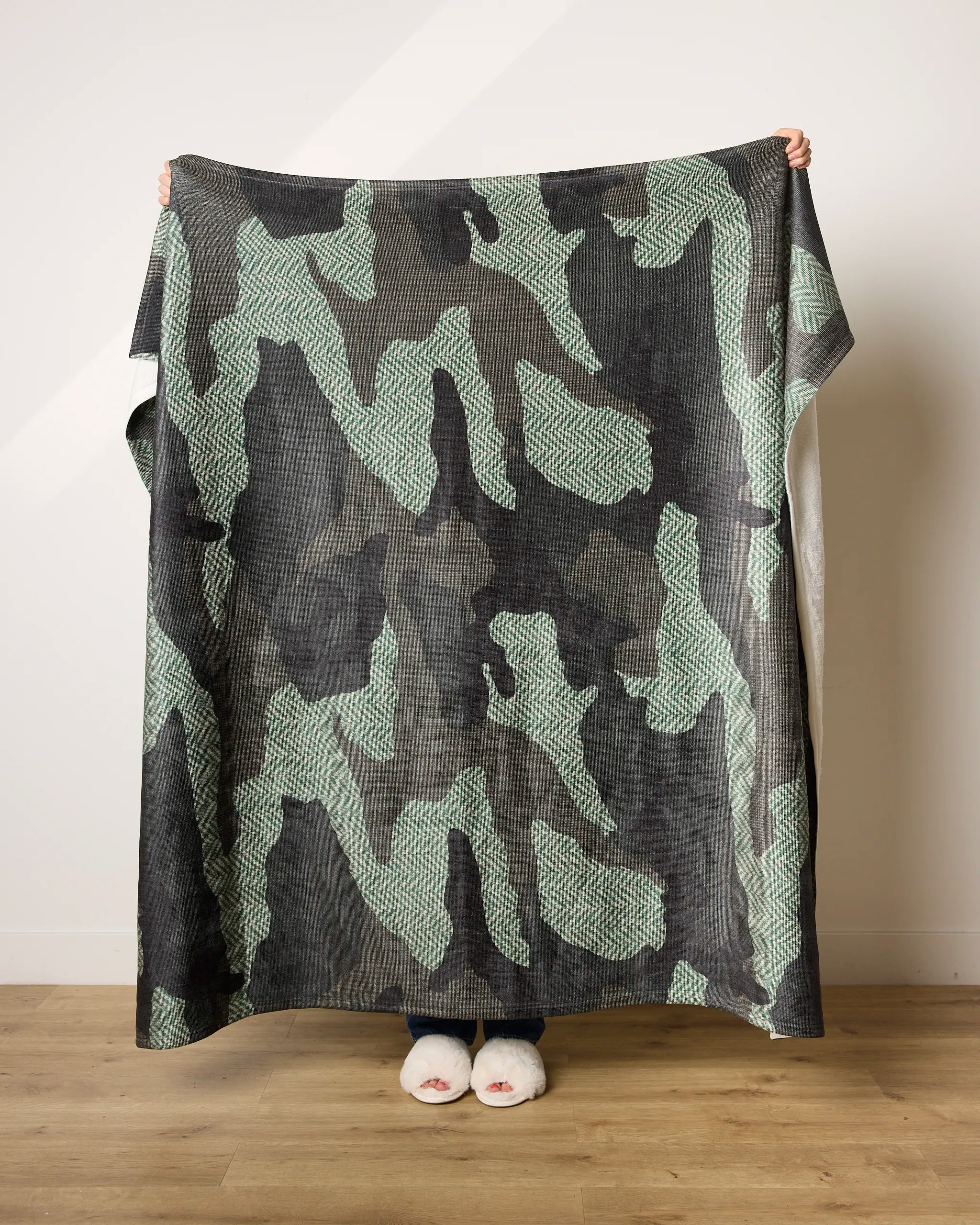 A person stands against a light wall, holding up the Calloway ultra-soft blanket by Geometry, featuring an abstract green and black pattern. Only their feet in white slippers and hands are visible.