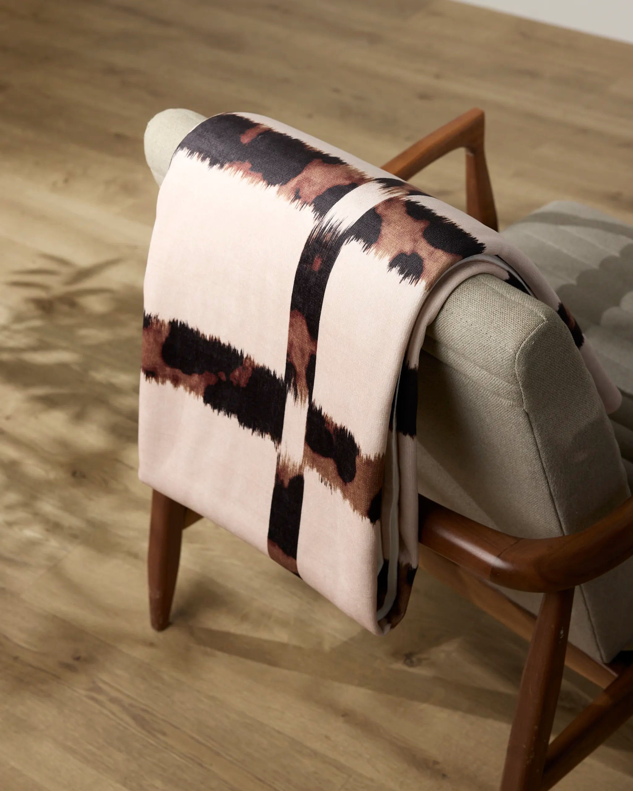 A Geometry Cheetah Grid blanket with a dark brown and black abstract stripe pattern is draped over the back of a light gray cushioned wooden chair on a light wood floor.