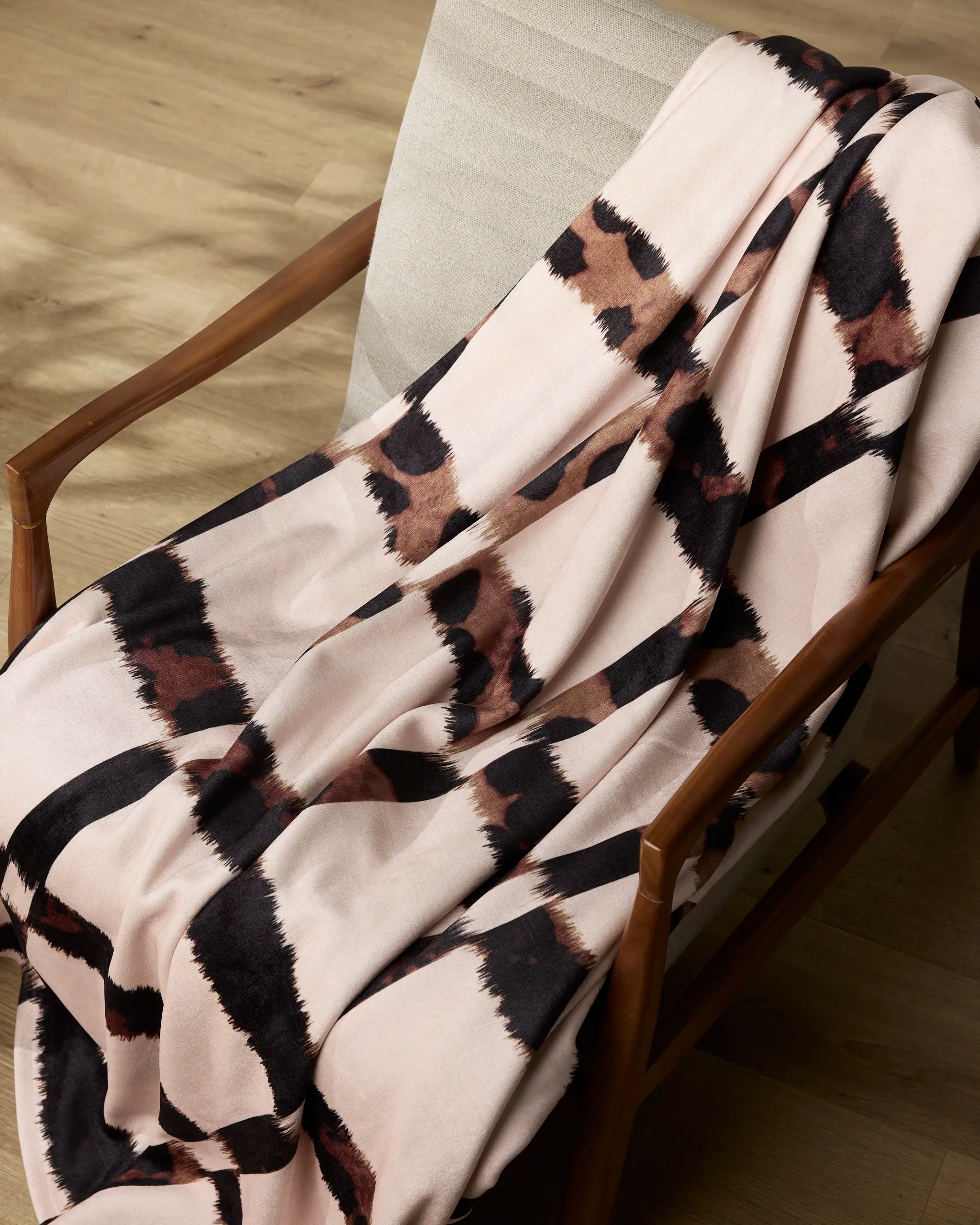 The Geometry Cheetah Grid Butter Blanket, featuring a bold abstract checkered pattern in black, brown, and tan, is draped over the arm and seat of a wooden chair with a light cushion on a wooden floor.