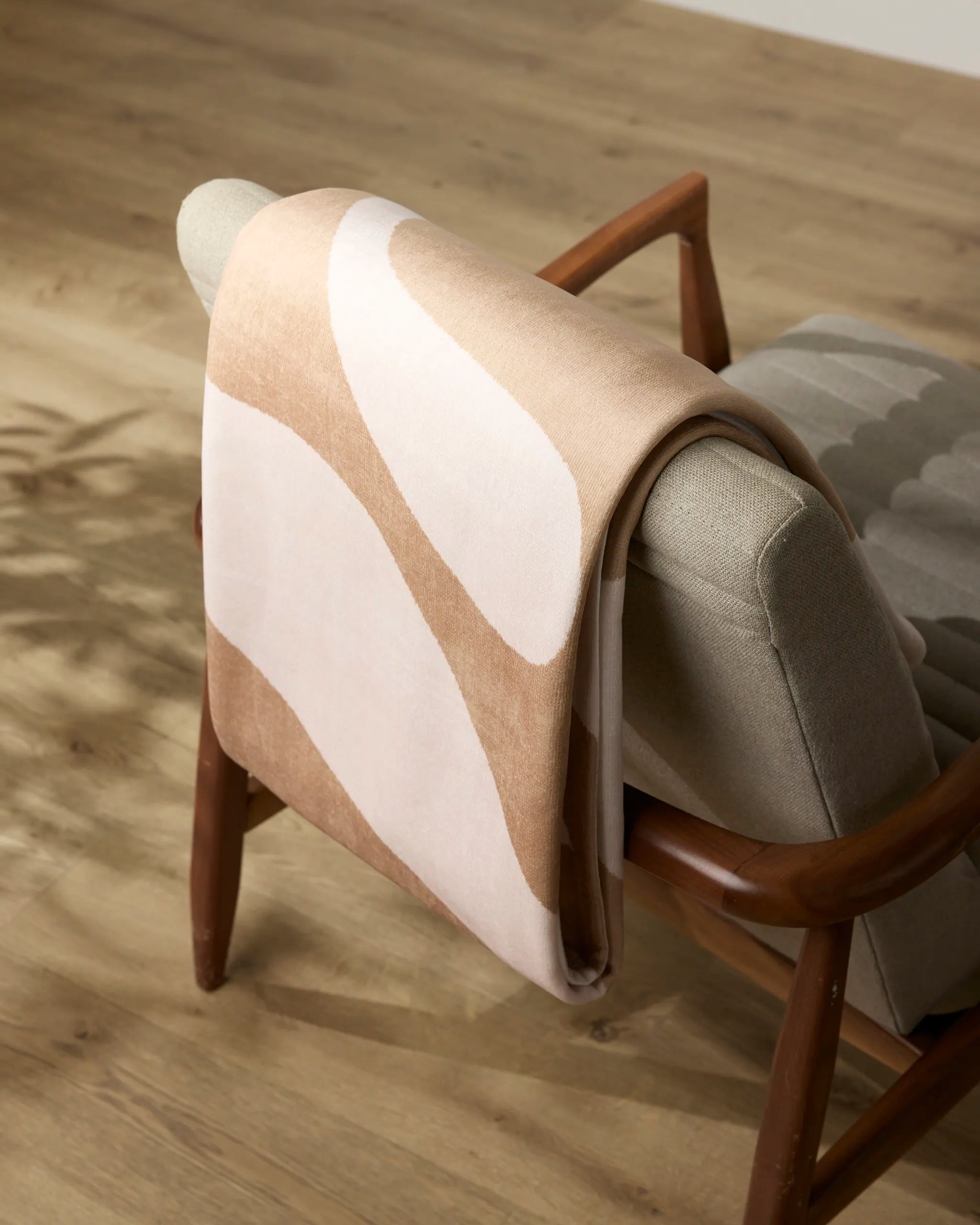 The Cleo throw by Geometry, featuring beige and light brown abstract patterns, is draped over the back of a mid-century modern armchair with light gray upholstery and wooden armrests on a light wood floor.