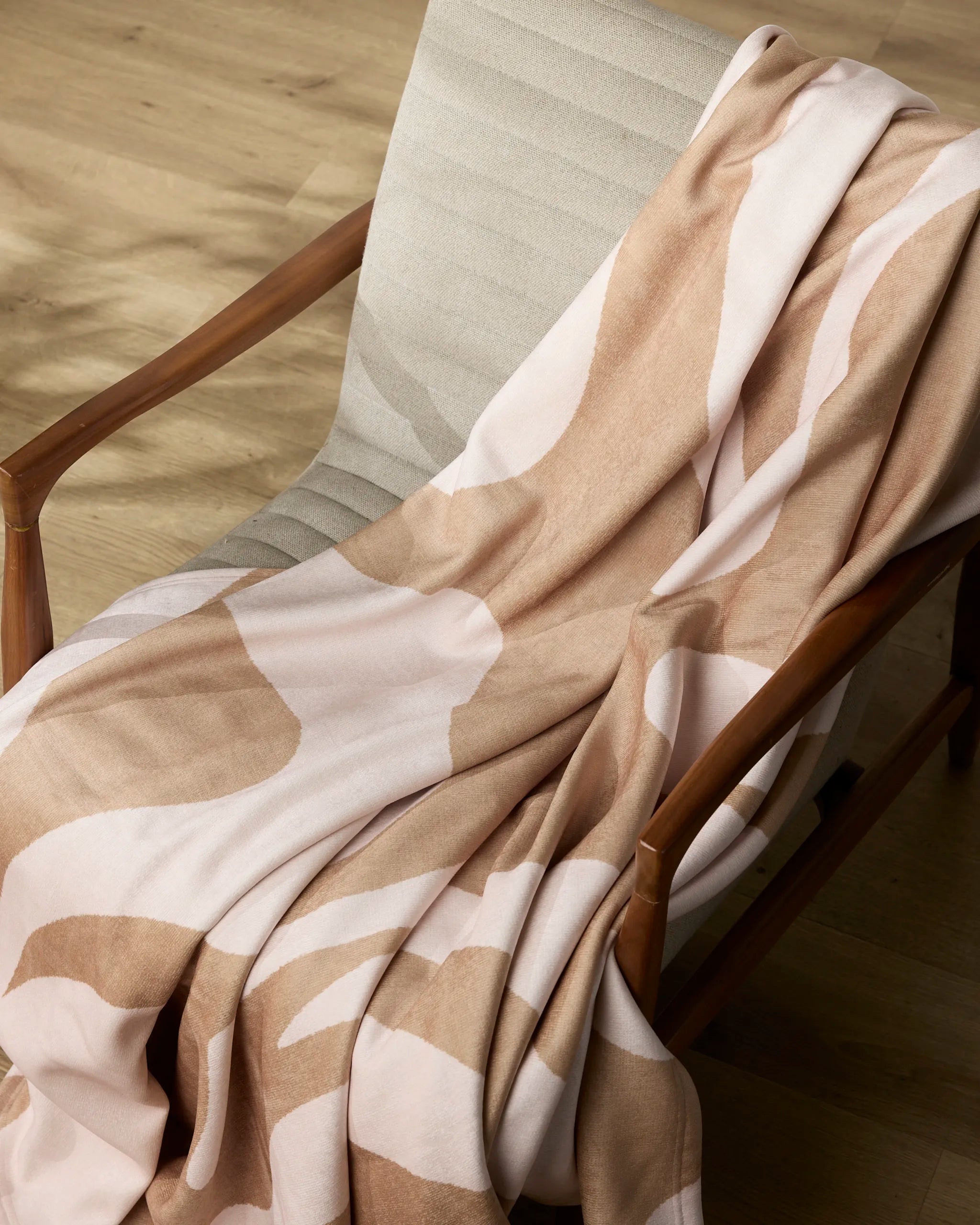 The Geometry Cleo recycled blanket, featuring a beige and white pattern, is draped over a light-gray cushioned wooden chair on a light wood floor.