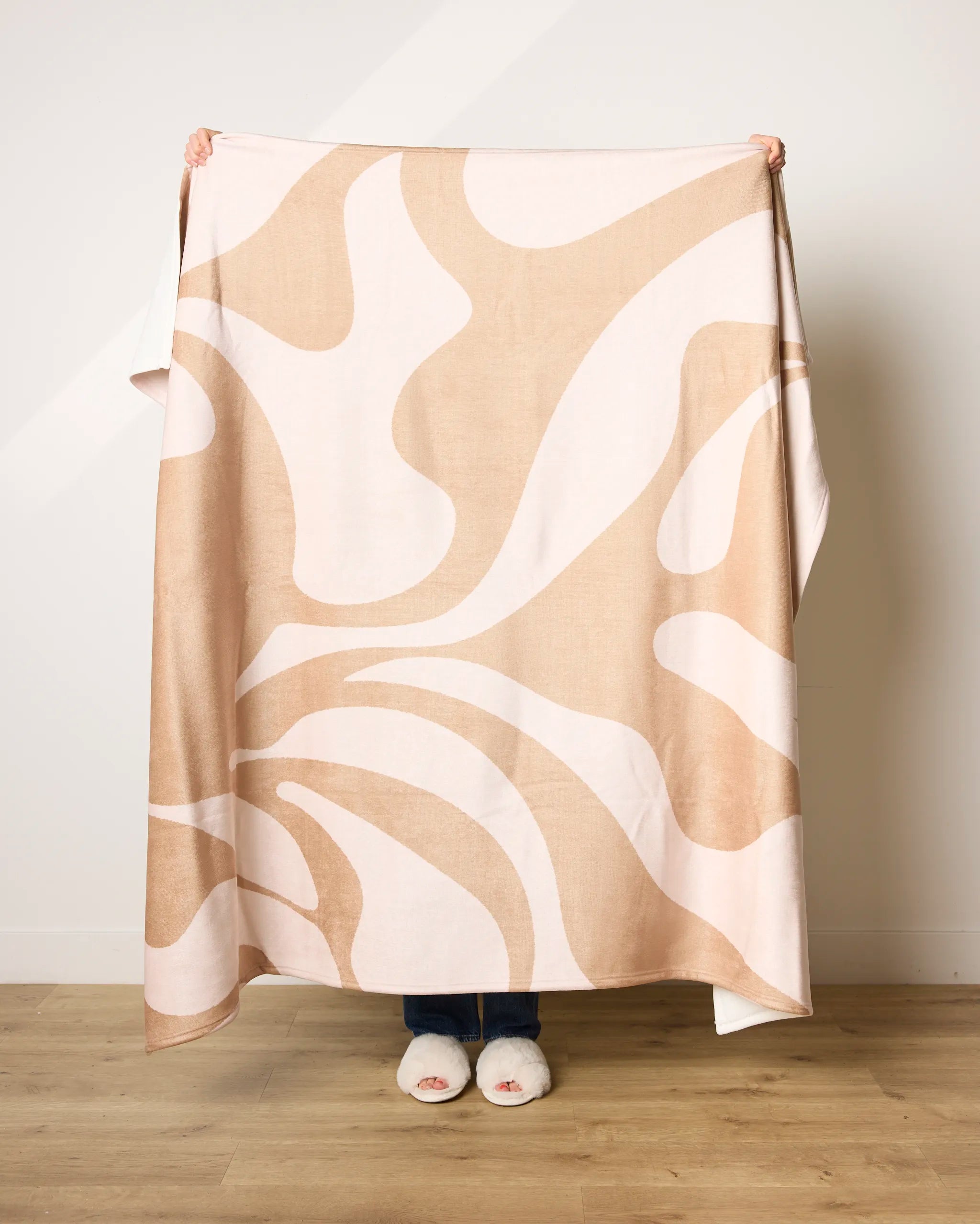 A person stands on a wooden floor holding up the Cleo blanket by Geometry, featuring ultra-soft beige and white abstract patterns crafted from recycled materials; only their slippered feet show below the blanket against a plain white wall.