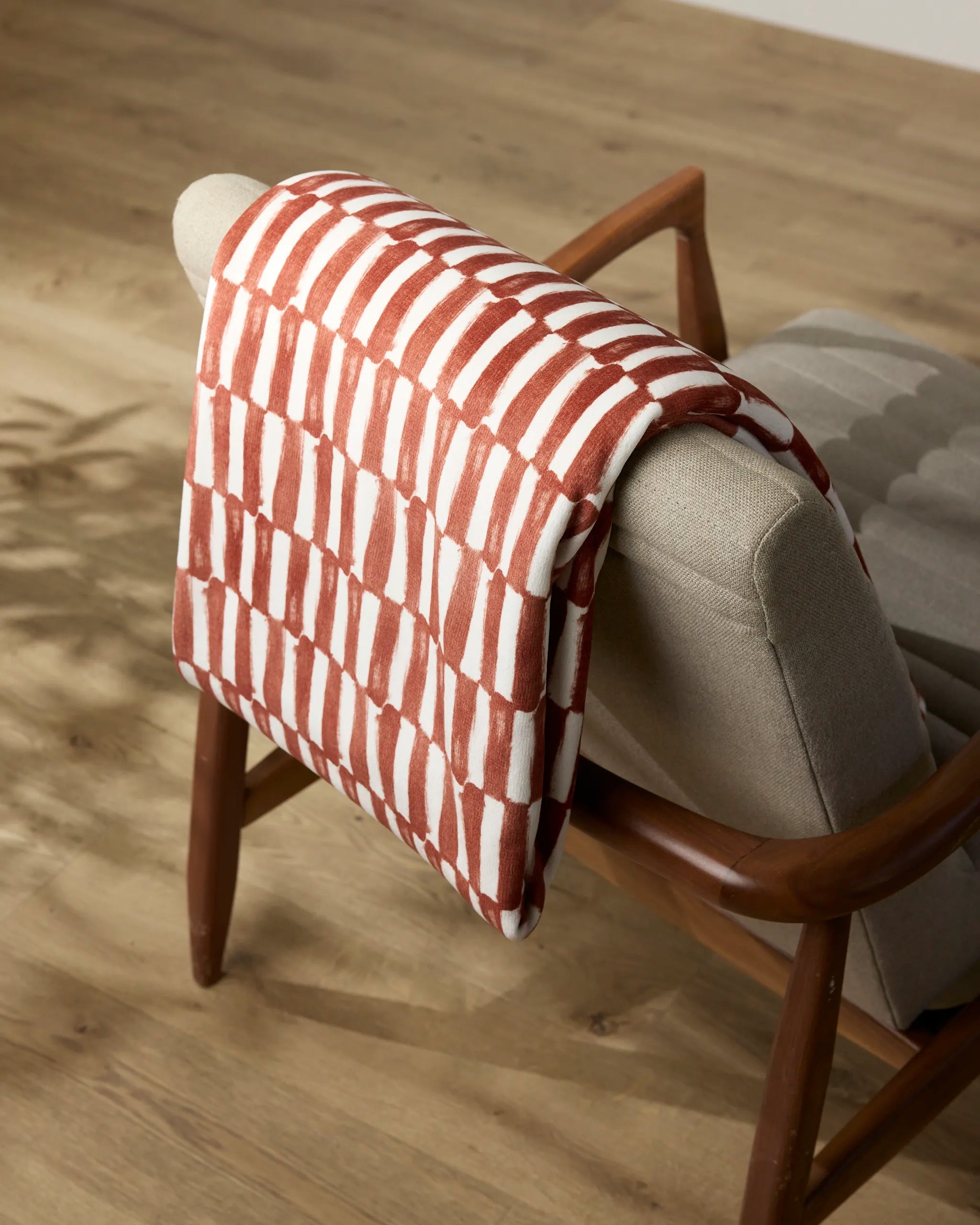 The Clove armchair by Geometry features beige upholstery, wooden arms and legs, and a soft silky throw draped over its back, resting on a light wood floor.