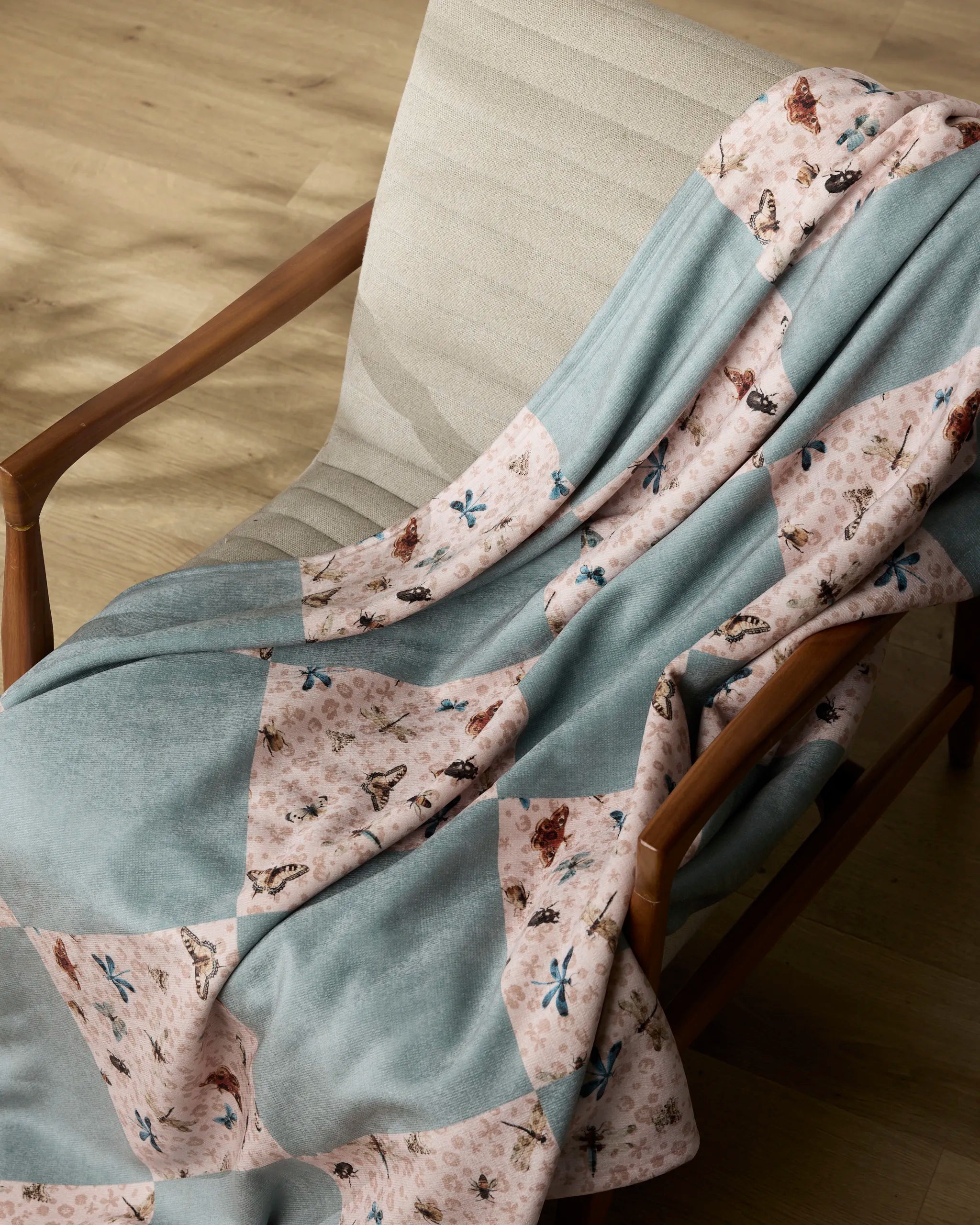 The Geometry Critter Check throw blanket, featuring alternating blue and pink floral-patterned squares, is draped over a light beige cushioned chair with wooden arms on a wooden floor.