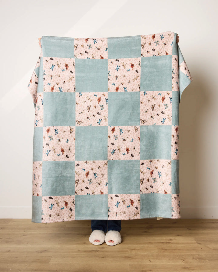 A person stands on a wooden floor holding up a Geometry Critter Check blanket, featuring blue and pink patchwork squares in ultra-soft fabric, covering their body with only their feet in white slippers showing.