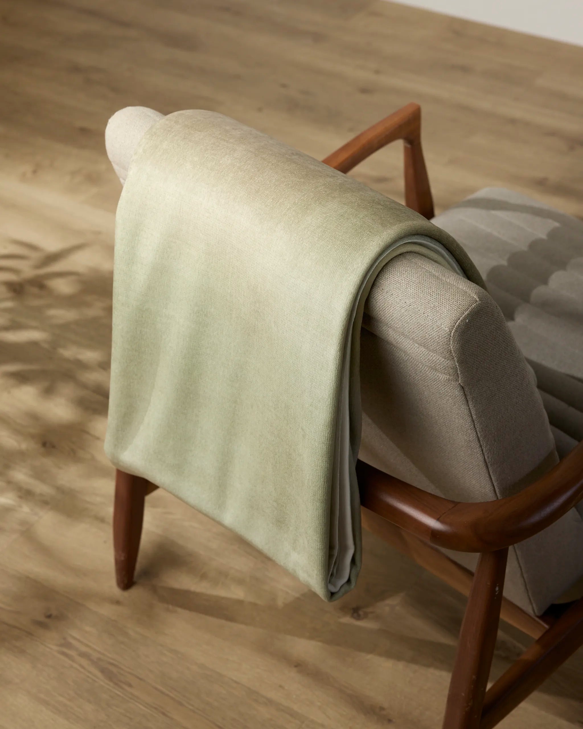The Ethereal throw blanket by Geometry is draped over a beige upholstered chair with wooden armrests, resting on a light wood floor with gentle shadows.
