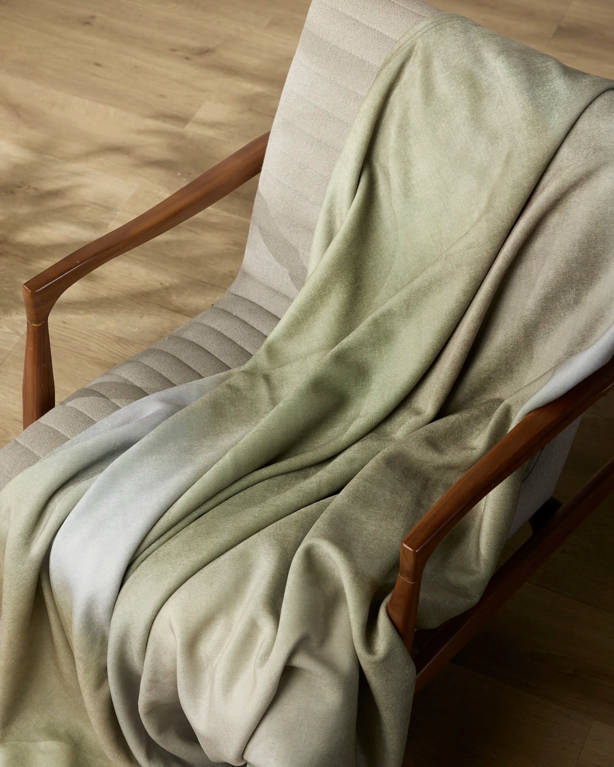 The Geometry Ethereal Butter Blanket, in a soft light green and beige gradient, is draped over a cushioned chair with wooden armrests on a light wood floor—a perfect cozy throw for relaxing evenings.