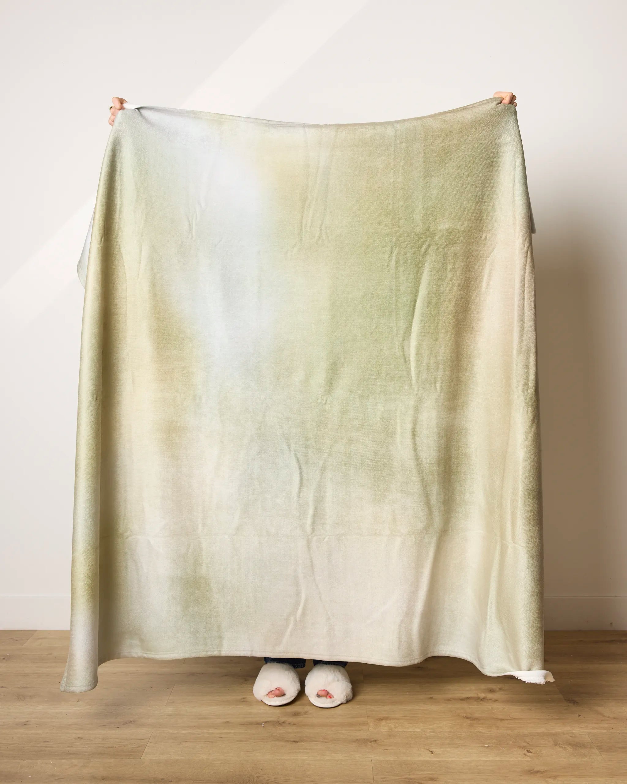 A person stands on a wooden floor holding up Geometry's Ethereal blanket, featuring a subtle light green and beige gradient. Only their hands and fuzzy-slippered feet are visible behind the ultra-soft fabric.