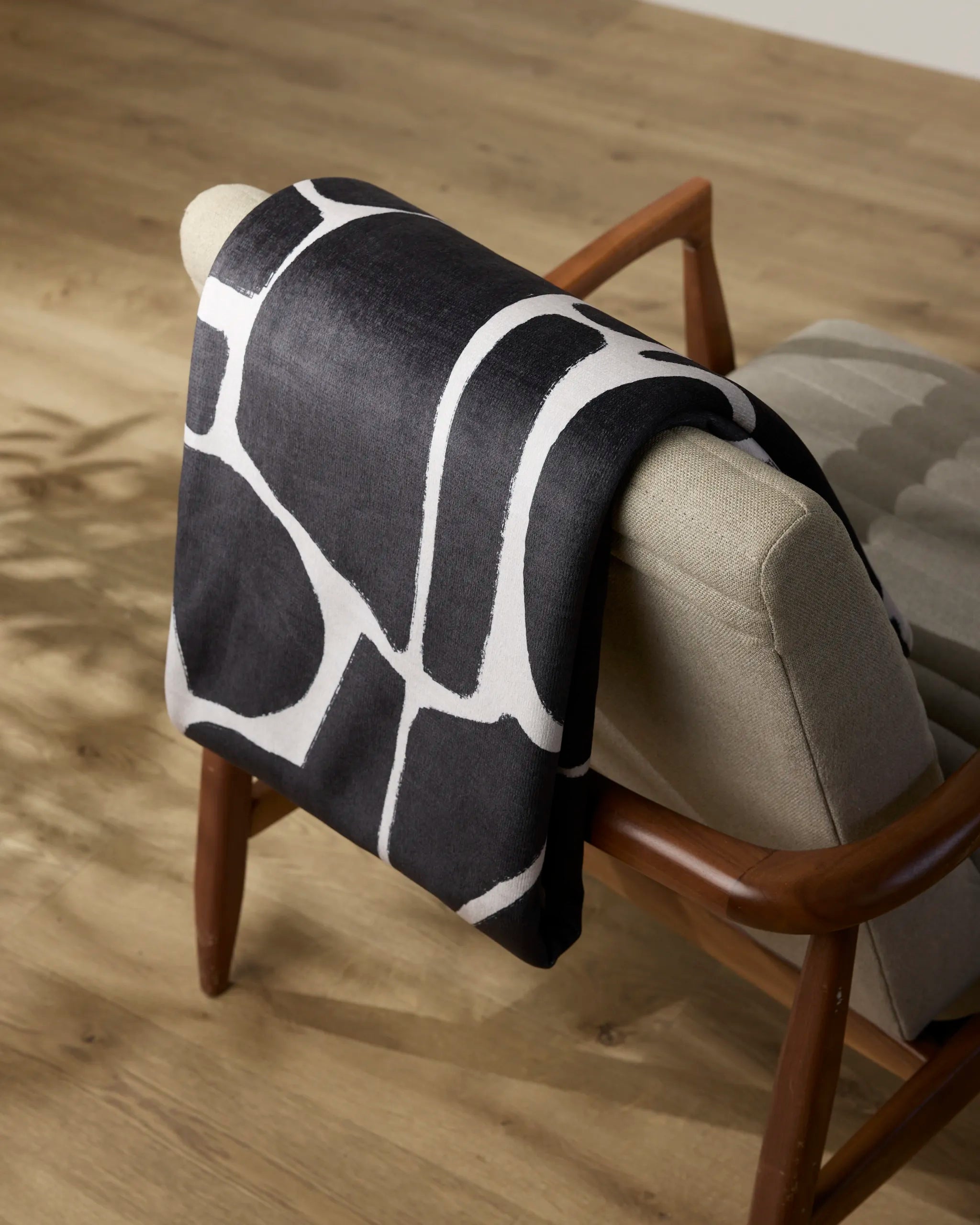 The Geometry Ezra throw blanket, featuring black and white abstract patterns, is draped over a light beige mid-century modern chair set on a wooden floor.