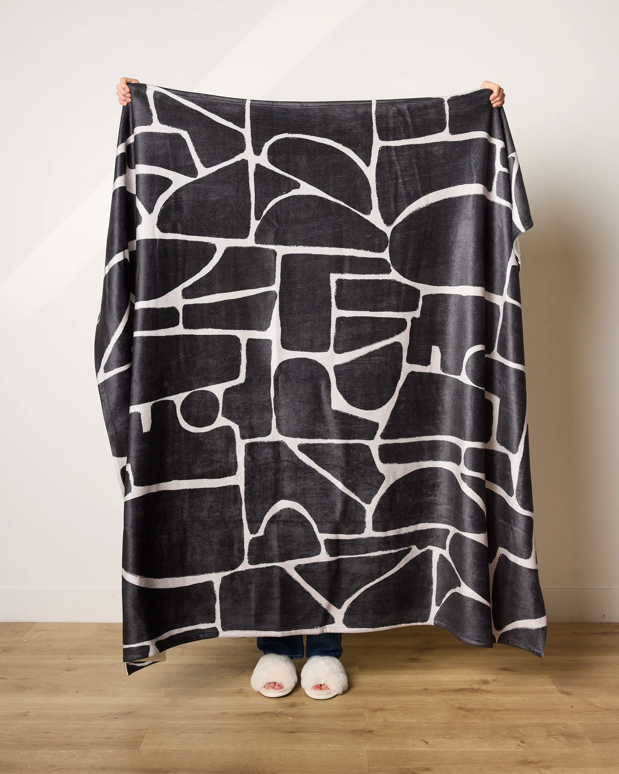 A person holds up the Ezra blanket by Geometry—an ultra-soft throw with an abstract geometric pattern. Only their hands and slippered feet are visible behind it, set against a plain white wall and wooden floor.