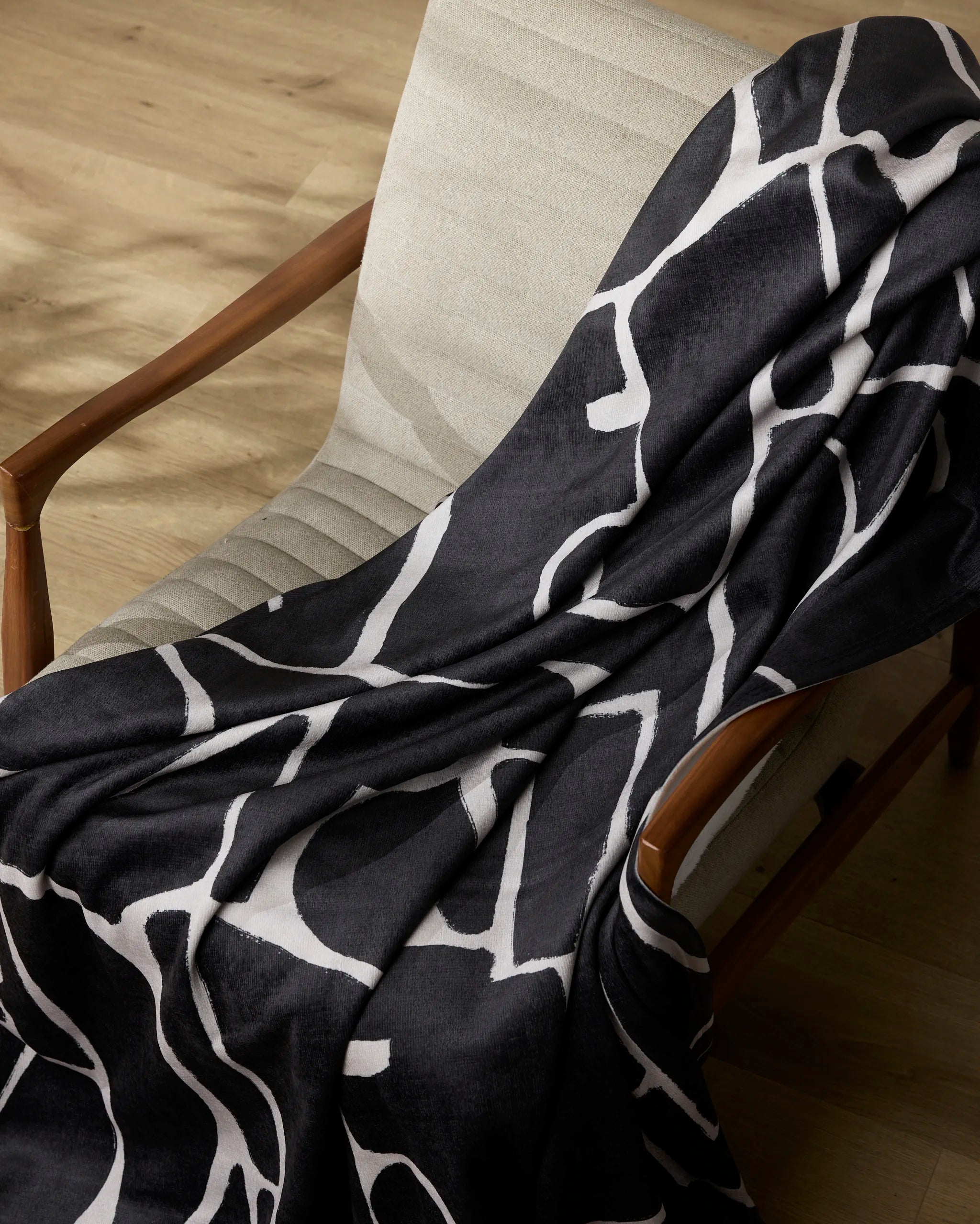The Ezra armchair by Geometry, featuring wooden arms and modern beige upholstery, has a bold white abstract-patterned throw draped over it and rests on a light wood floor.