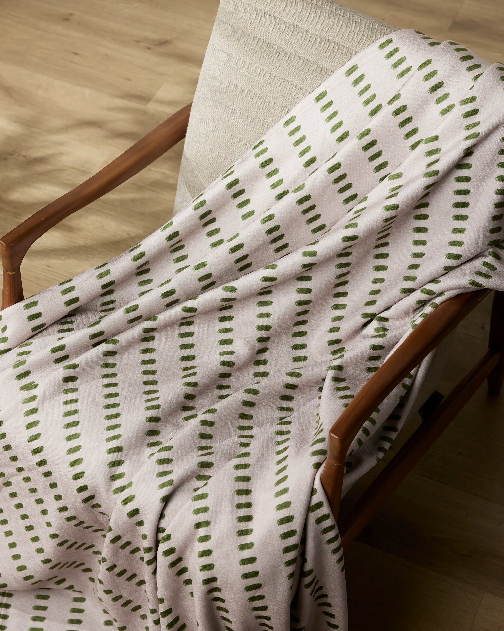 The Gracie throw blanket by Geometry, featuring green dashed lines, adds comfort and style as it drapes over a wooden armchair with a beige cushion on a wooden floor.
