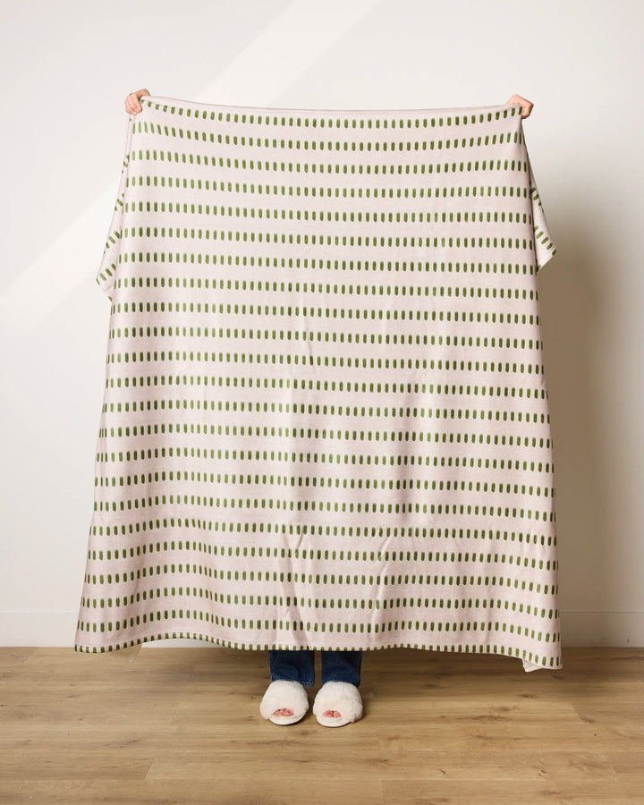 A person indoors holds up Geometry’s Gracie ultra-soft blanket, made from recycled materials and featuring green vertical lines; only their hands, slippered feet, and blue jeans are visible.