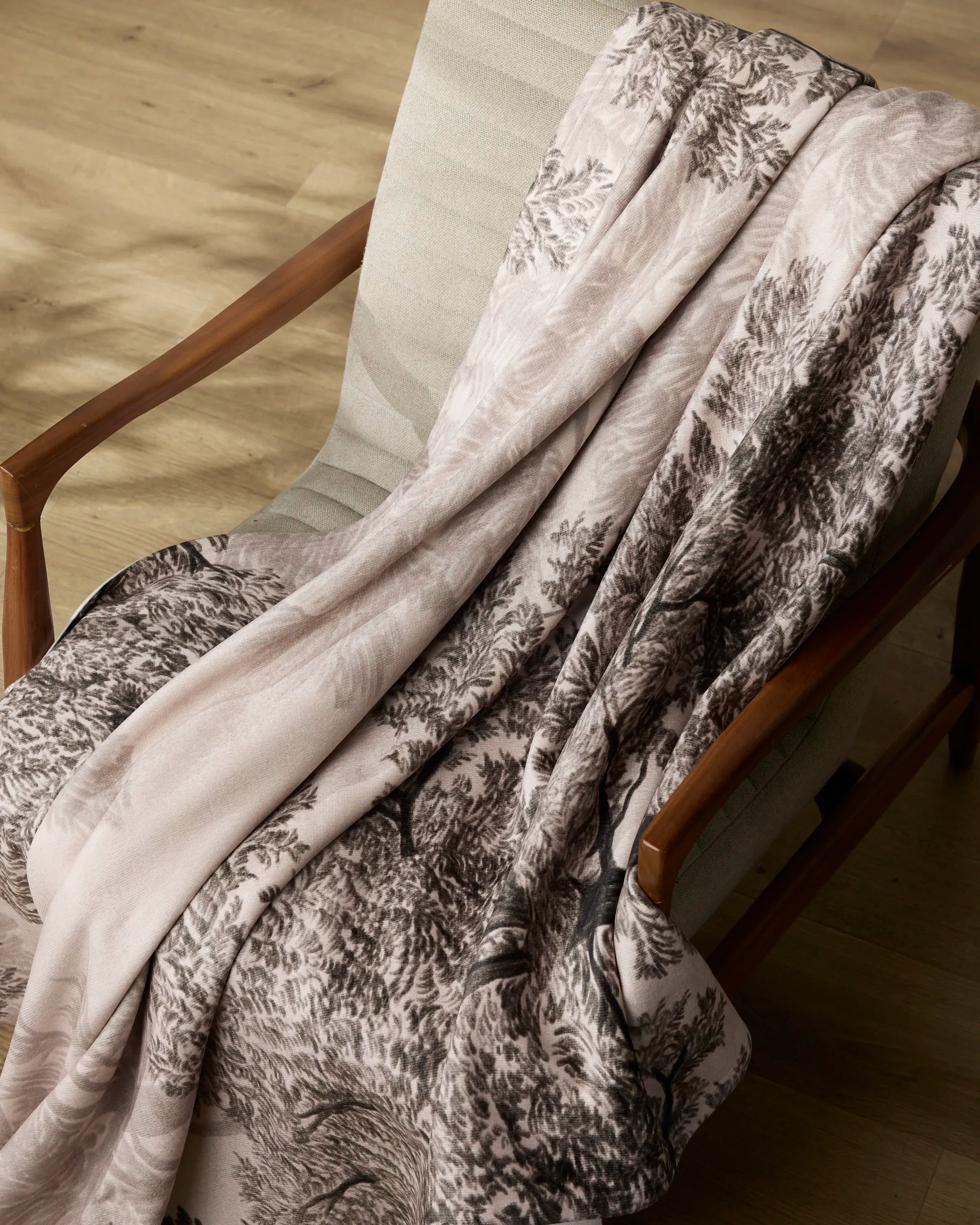 The Geometry Haven blanket, made from recycled materials and featuring a nature-inspired pattern, is draped over a light upholstered chair with wooden armrests on a wooden floor.