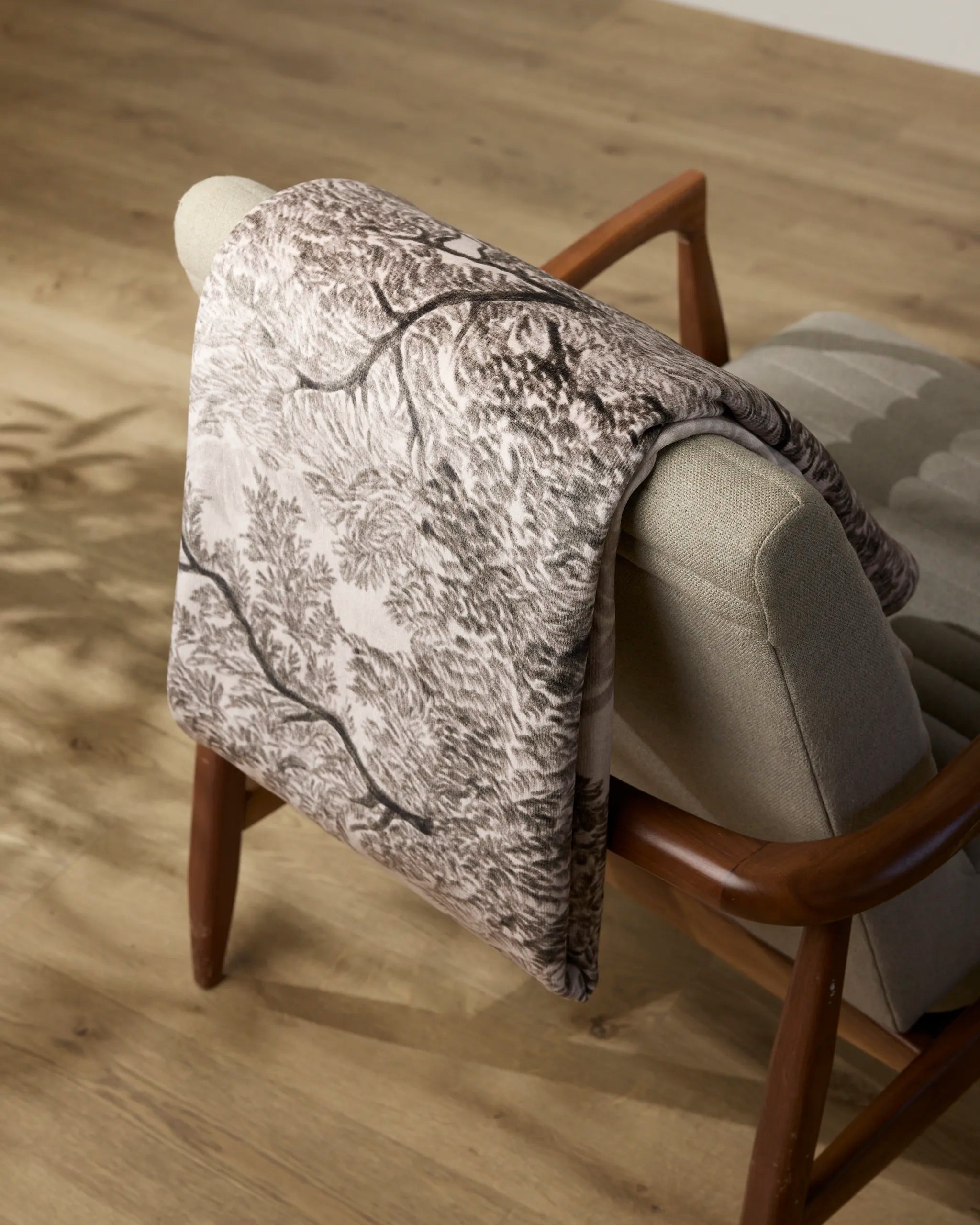 The Geometry Haven blanket, featuring tree and foliage patterns, is draped over a light grey upholstered chair with wooden arms on a light wood floor.