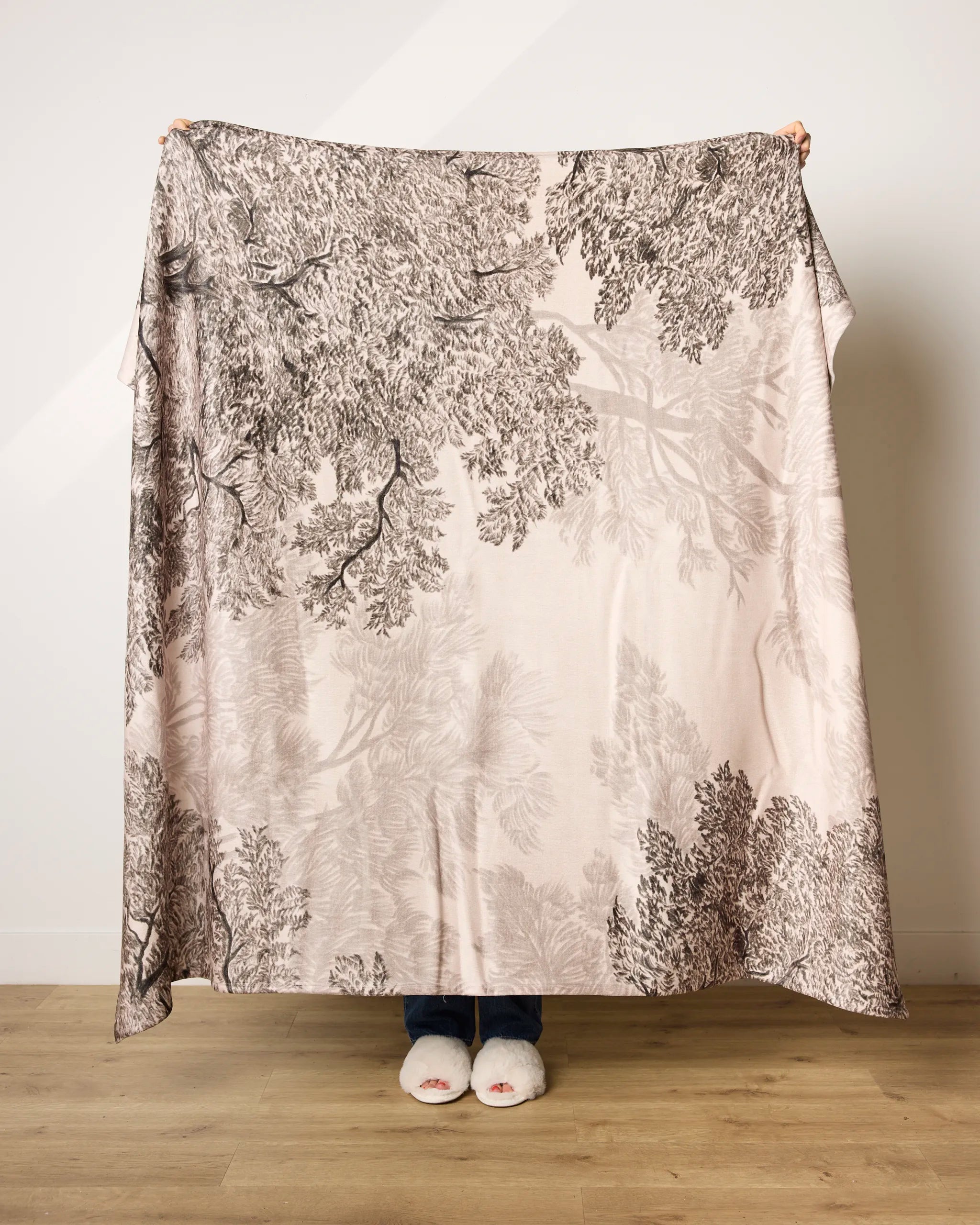 A person stands indoors holding up Geometry's Haven blanket with a black and white tree branch pattern; only their hands, slipper-clad feet, and toes peek out behind the ultra-soft fabric made from recycled materials.