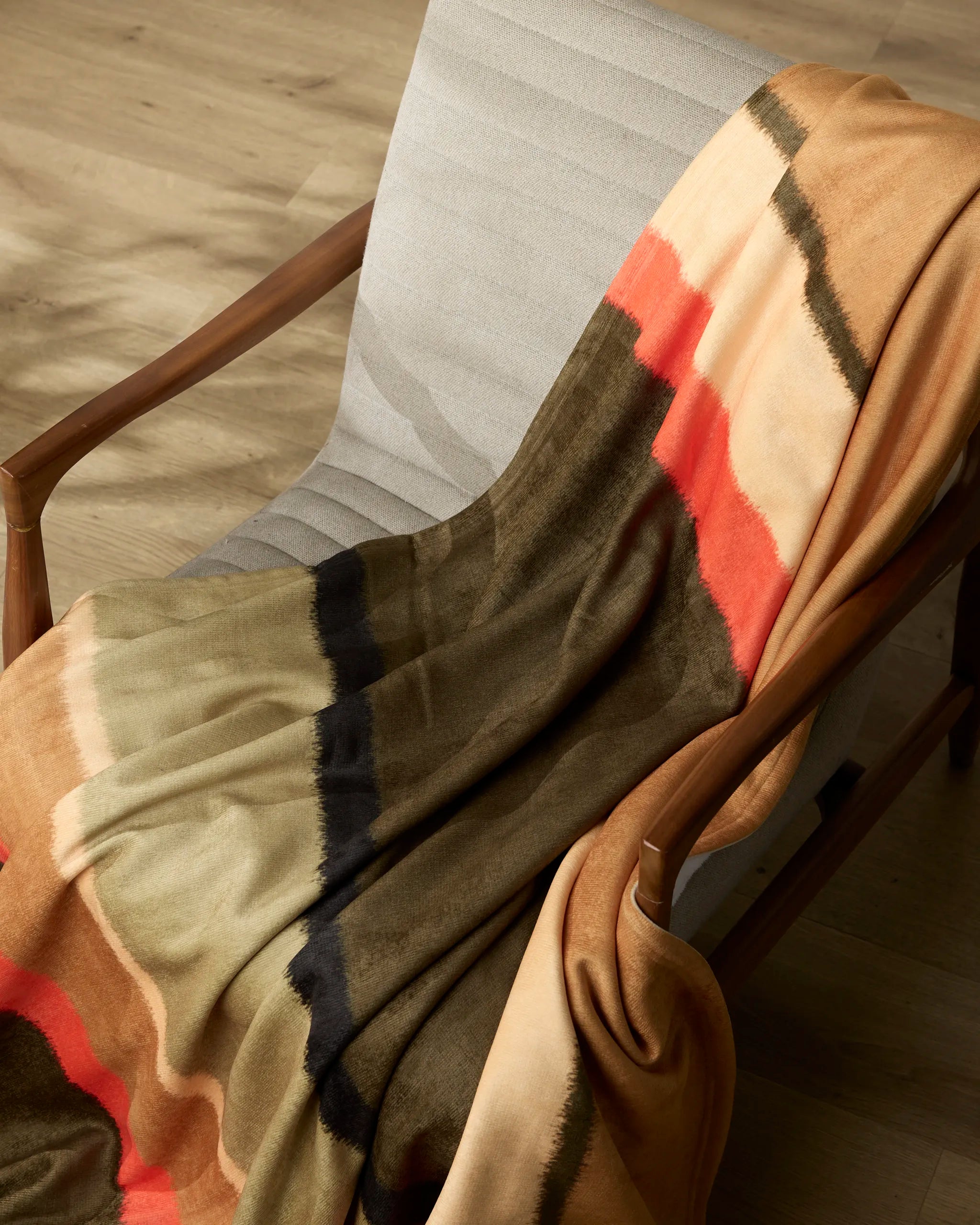 The Geometry Hunting Lodge blanket, featuring beige, olive, black, and orange stripes, is draped over a light gray cushioned chair with wooden armrests on a wooden floor.