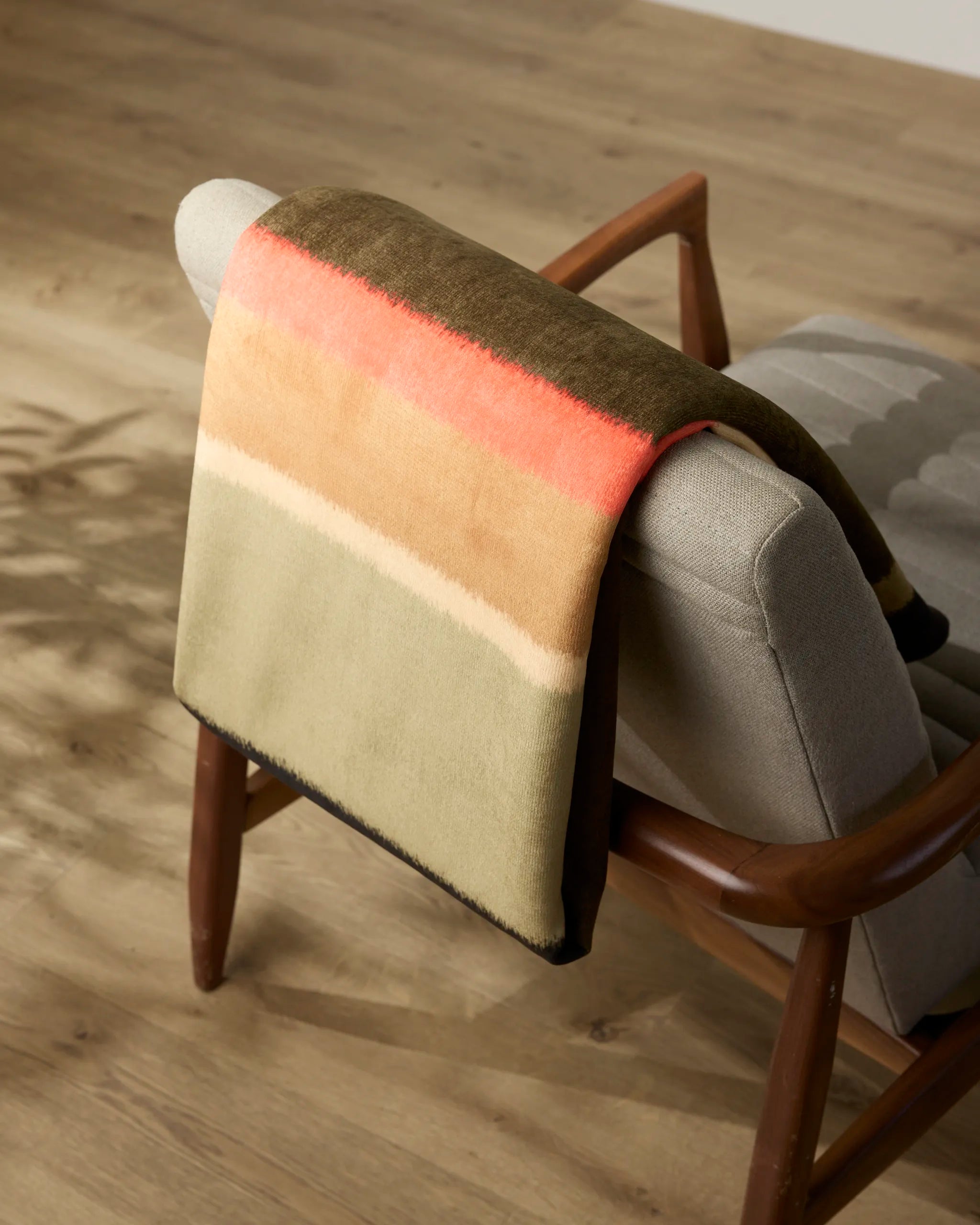 A folded Hunting Lodge blanket by Geometry, featuring earthy tones and a coral stripe, is draped over the back of a light gray upholstered chair with wooden arms on a light wood floor.