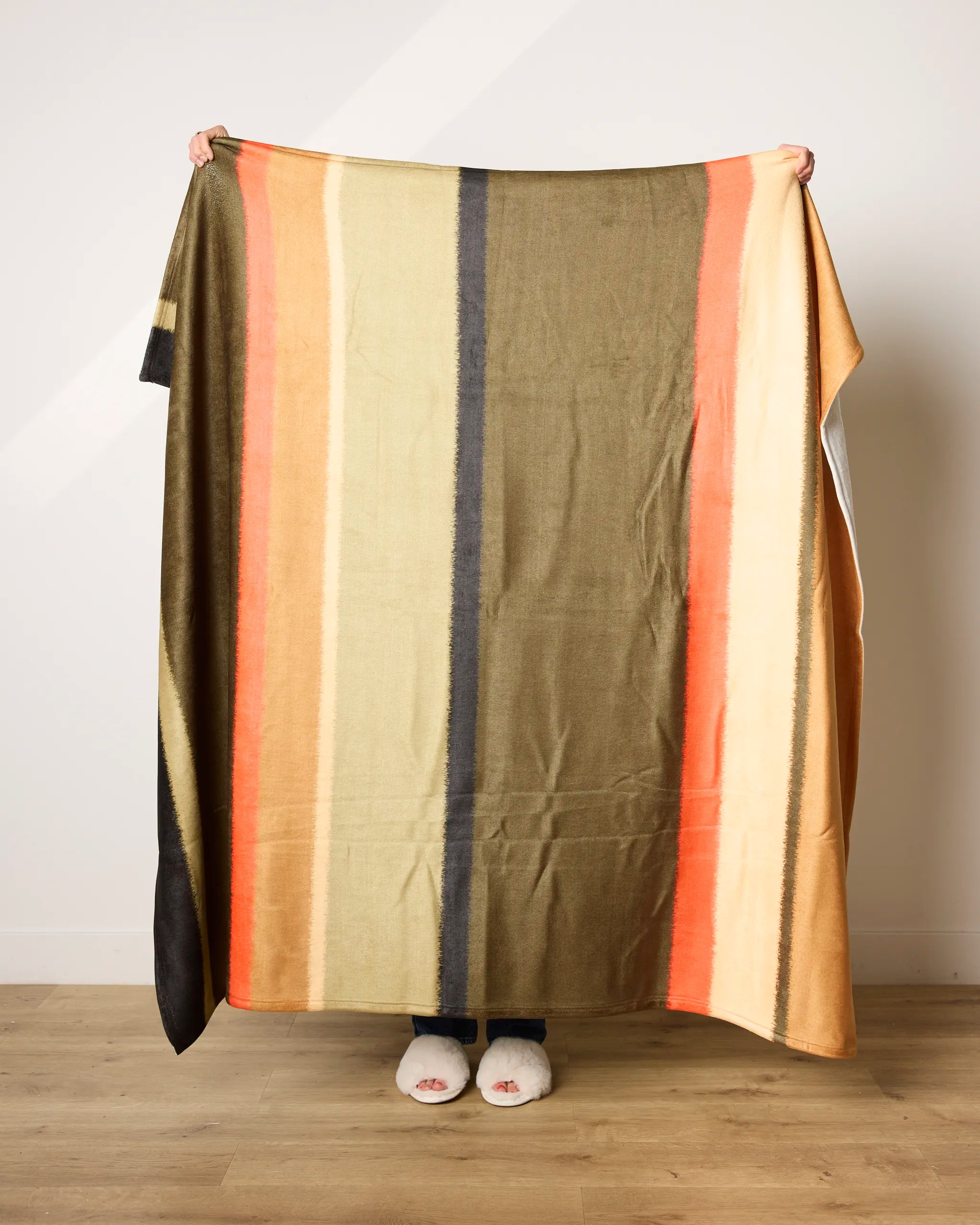 A person stands indoors holding up the Geometry Hunting Lodge blanket with vertical stripes in olive green, orange, beige, and black. Only their hands and feet in white slippers are visible. This ultra-soft blanket is made from recycled materials.