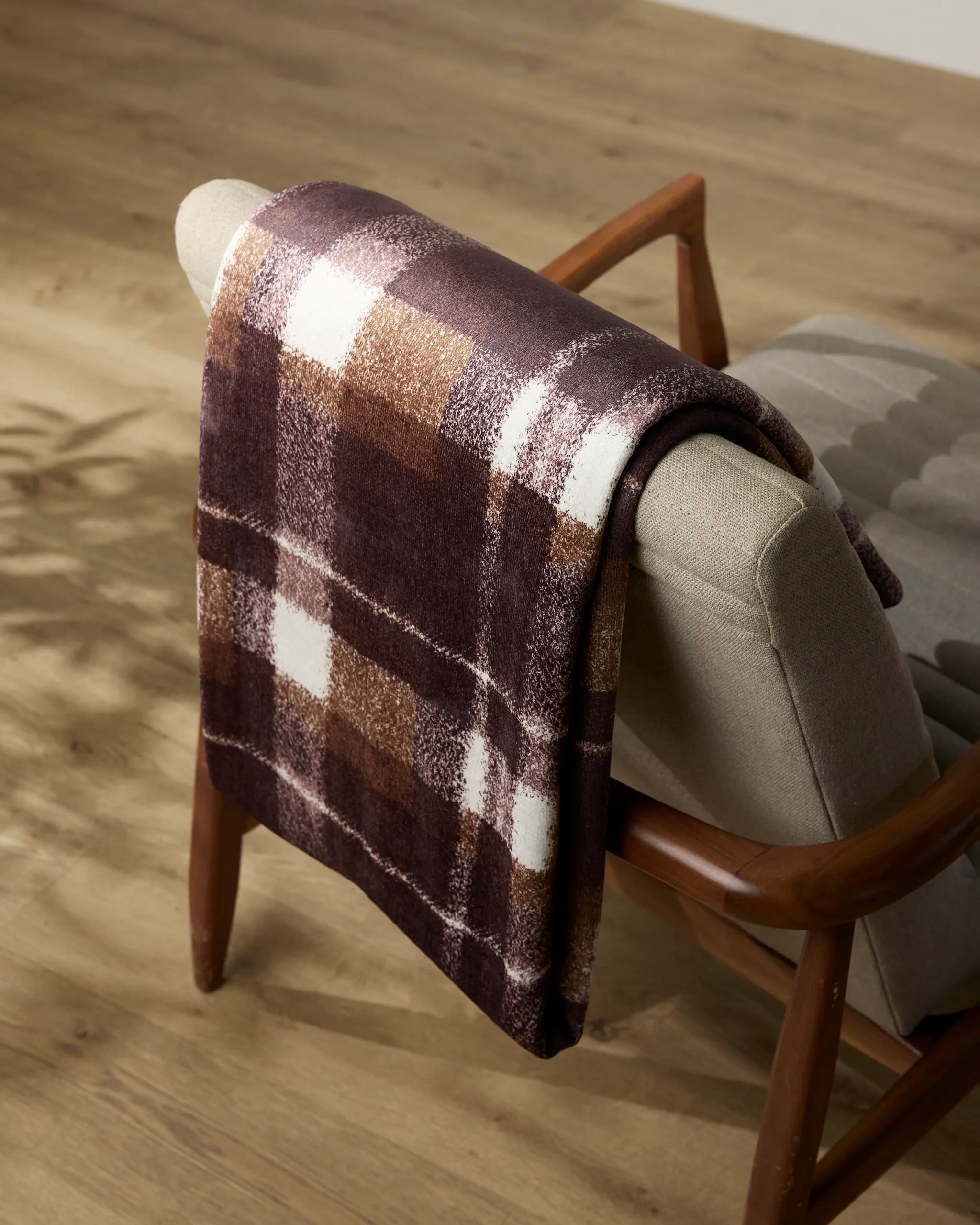 The Geometry Lennon throw blanket in brown and white plaid is draped over a light gray upholstered chair with wooden arms, set on a light wood floor—a cozy accent for any room.