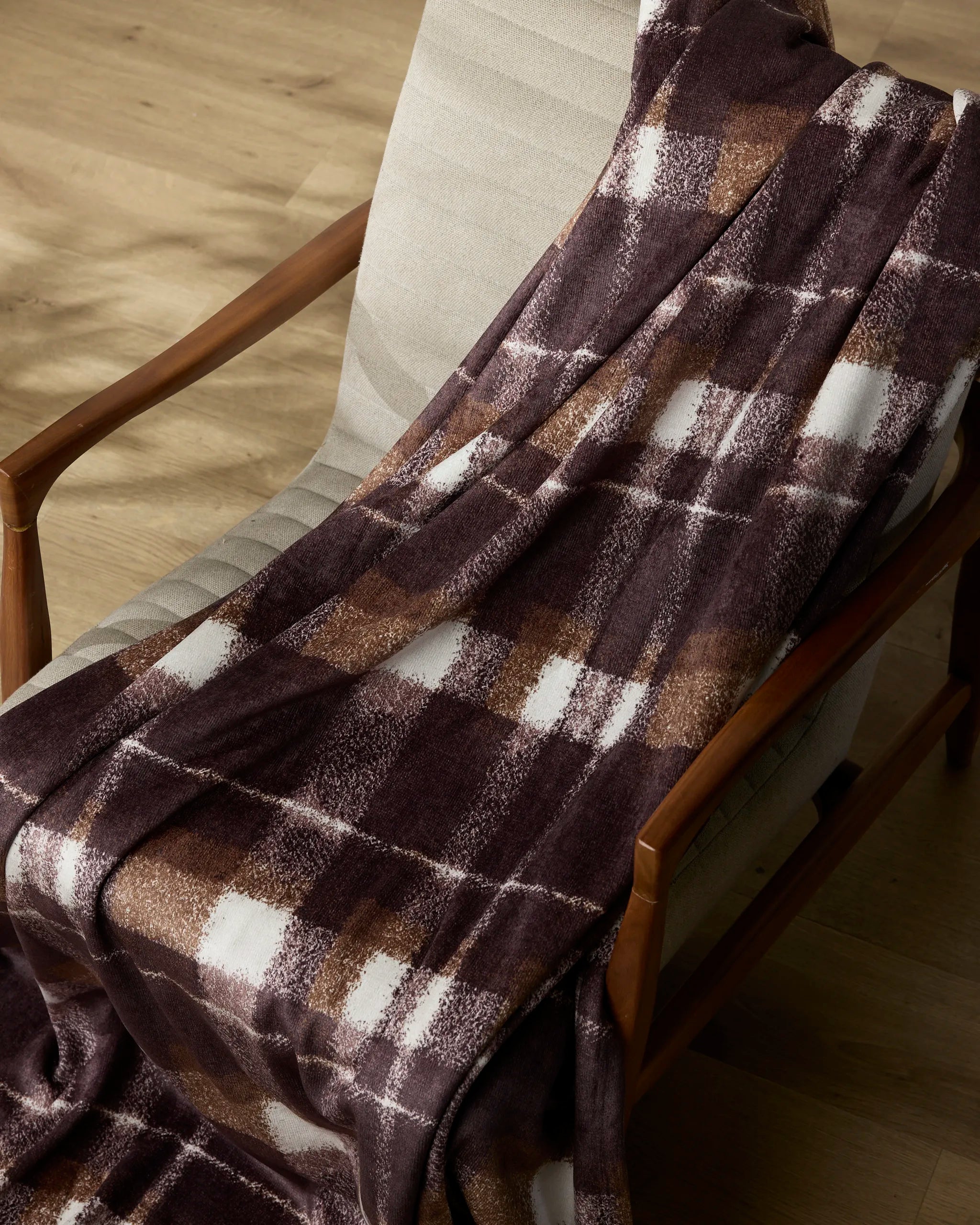 The Geometry Lennon throw, in brown and white plaid, is draped over a light beige upholstered wooden chair on a light wood floor.