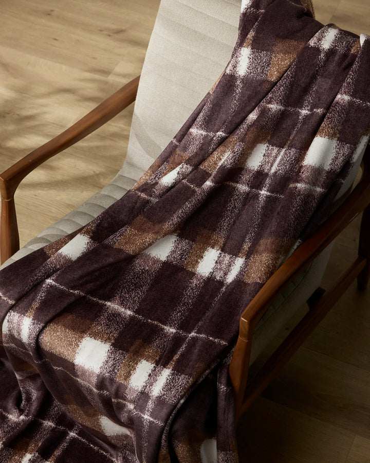 The Geometry Lennon throw, in brown and white plaid, is draped over a light beige upholstered wooden chair on a light wood floor.