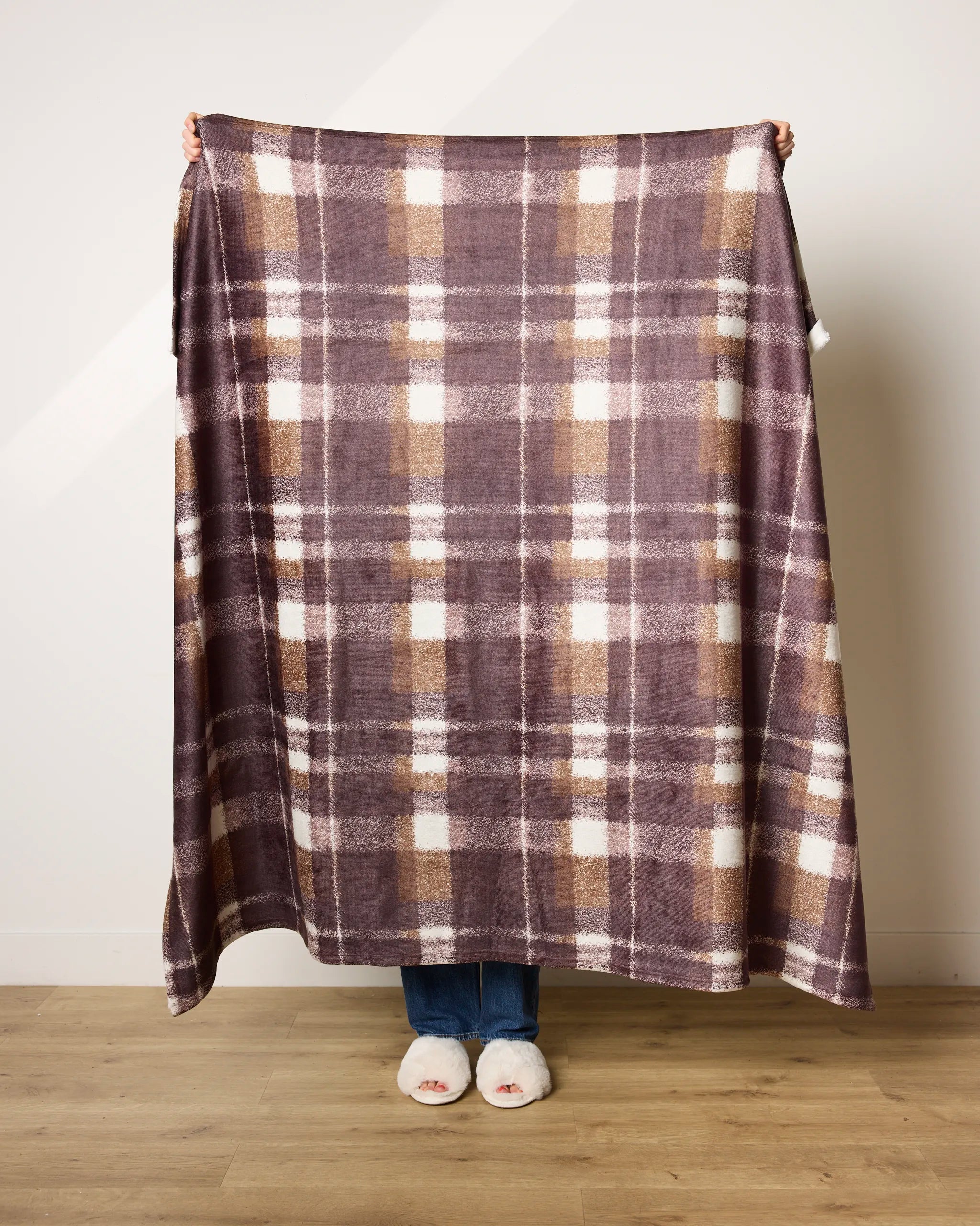 A person holds up the Lennon blanket by Geometry, featuring a large brown and beige plaid design. Made from recycled materials, the ultra-soft blanket drapes to reveal white slippers on a wooden floor.