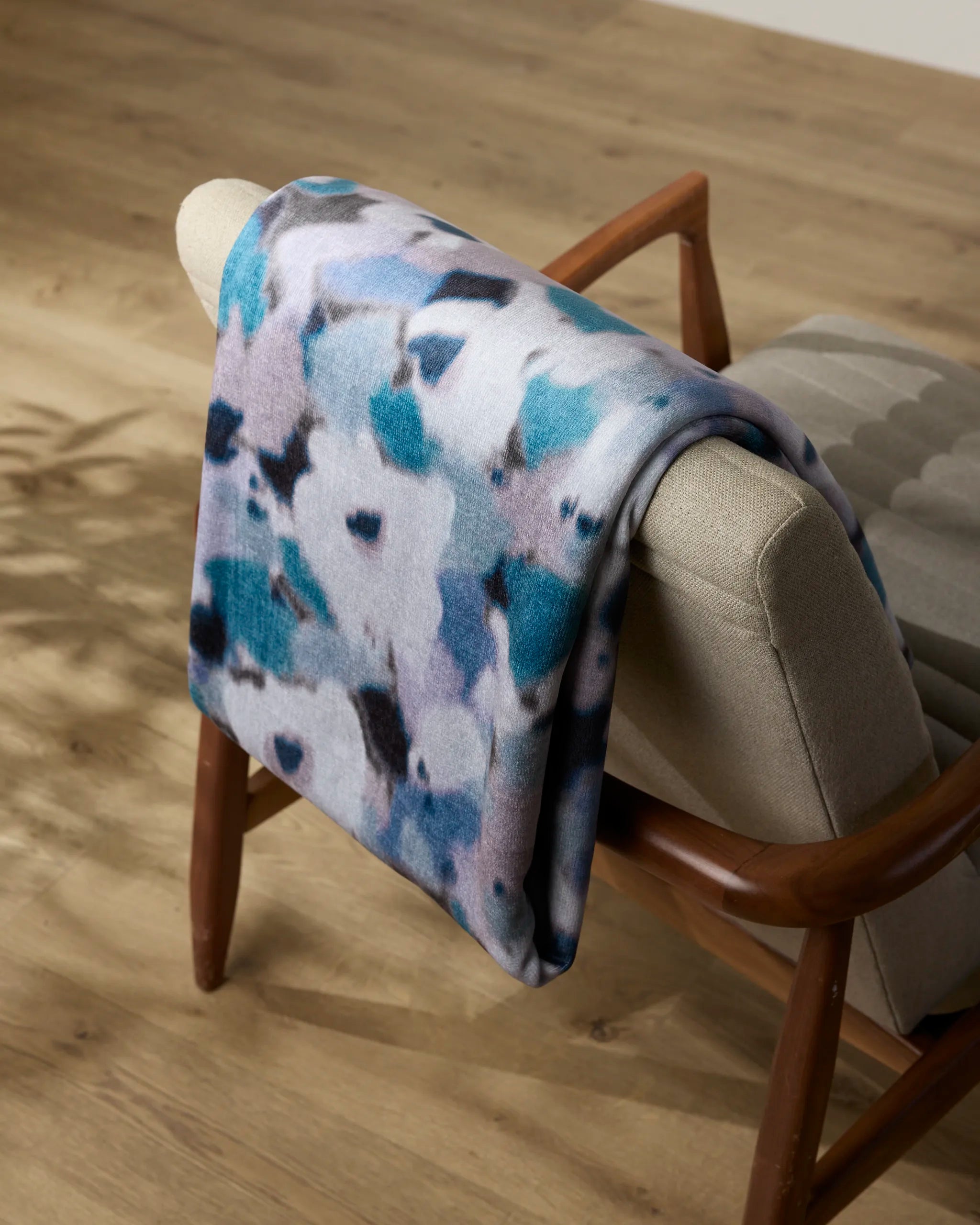 The Geometry Muted Bloom throw blanket, with blue, purple, black, and white patterns, is draped over a light beige mid-century modern armchair on a wooden floor.