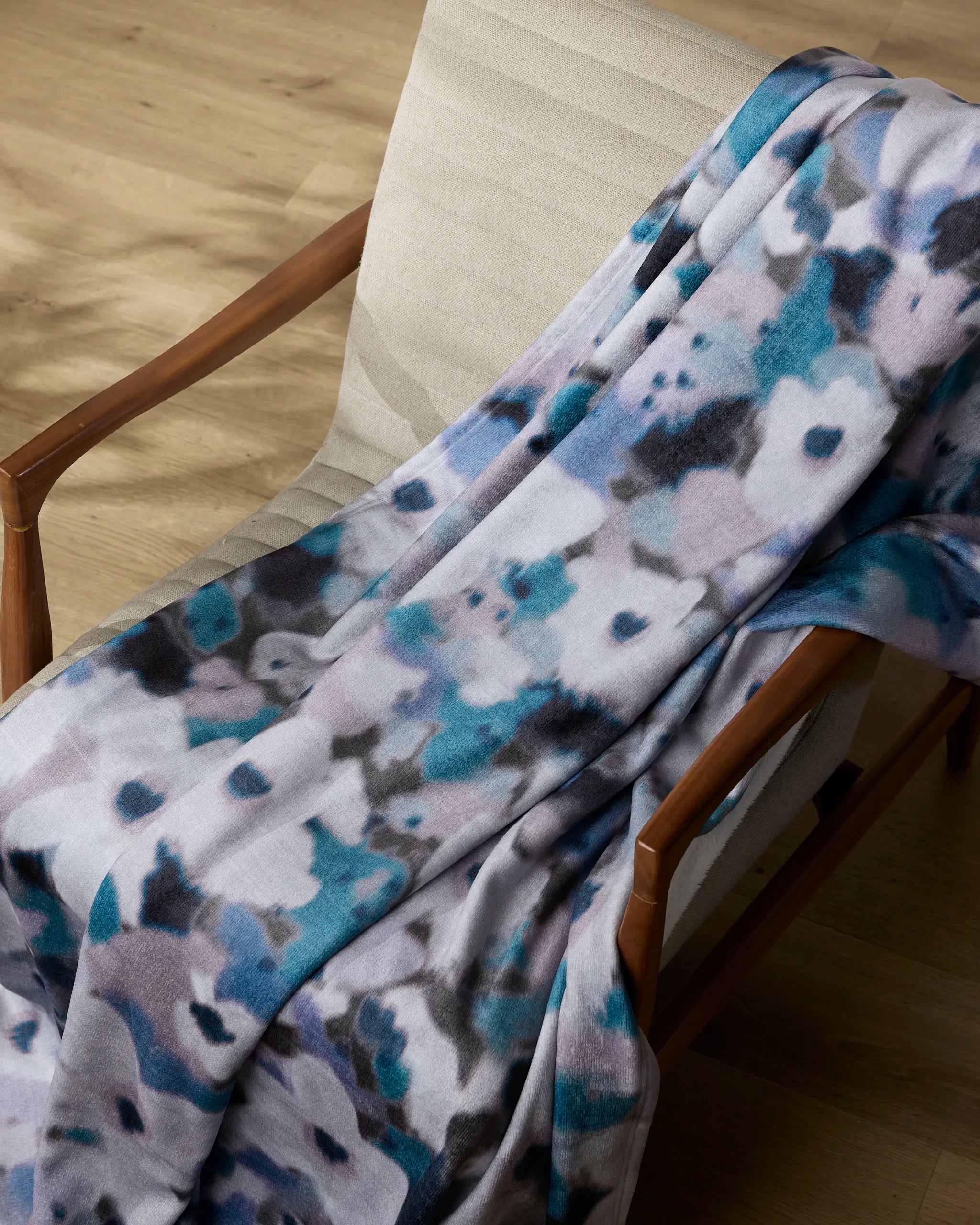 The Geometry Muted Bloom throw blanket, with its soft fabric and abstract blue, gray, and black pattern, is draped over a light padded chair with wooden armrests on a wooden floor.
