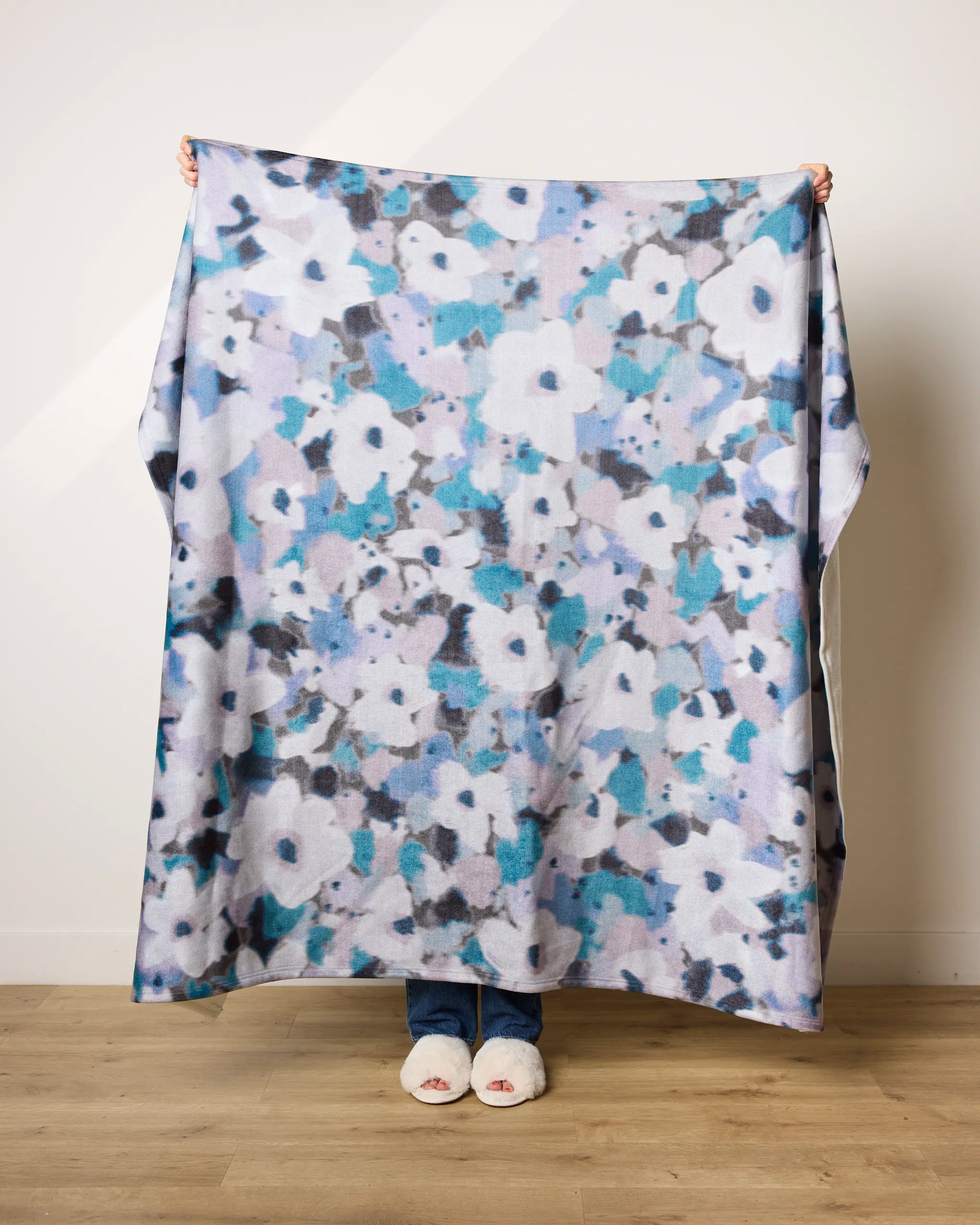 A person indoors on a wooden floor holds up the Geometry Muted Bloom ultra-soft blanket, featuring a blue, white, and gray abstract floral pattern. Only their feet in white slippers and hands are visible. Made from recycled materials.
